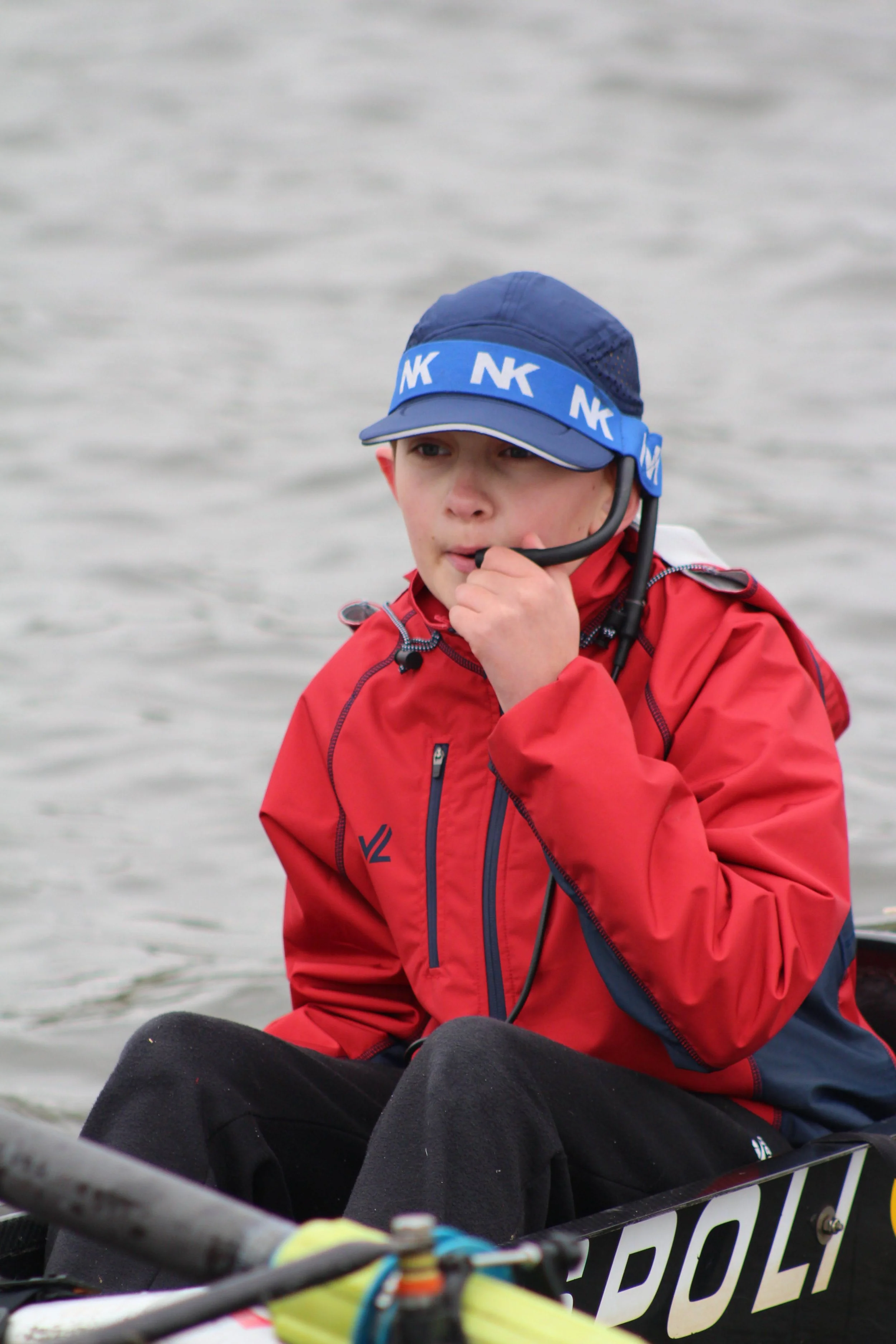 A young boy in a red jacket and blue cap with 'NK' logos, sitting in a boat on a body of water, holding a communication device to his mouth.