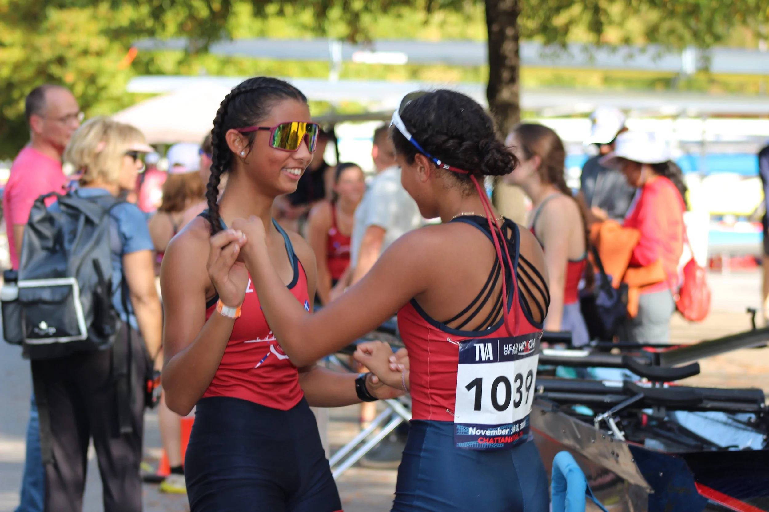 Two female Elizabeth City Rowing Club athletes in red uniforms are smiling and shaking hands at a competitive rowing event, with spectators in the background.