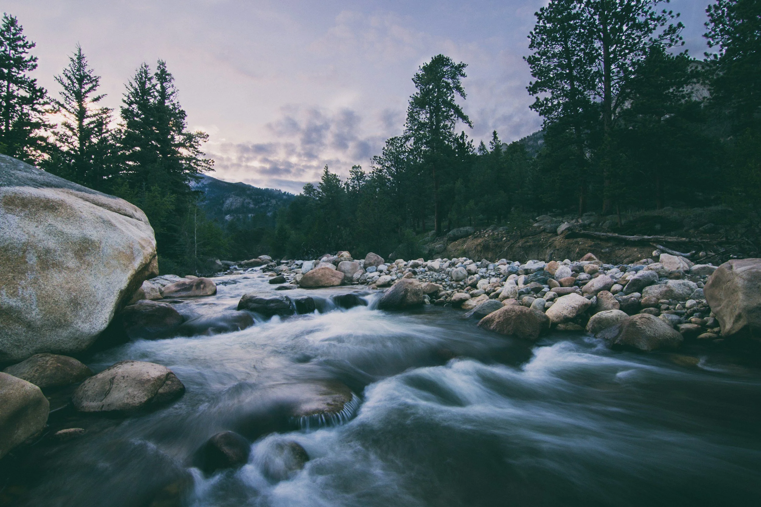 A flowing mountain stream surrounded by rocks and trees, with a sky filled with clouds in the background.