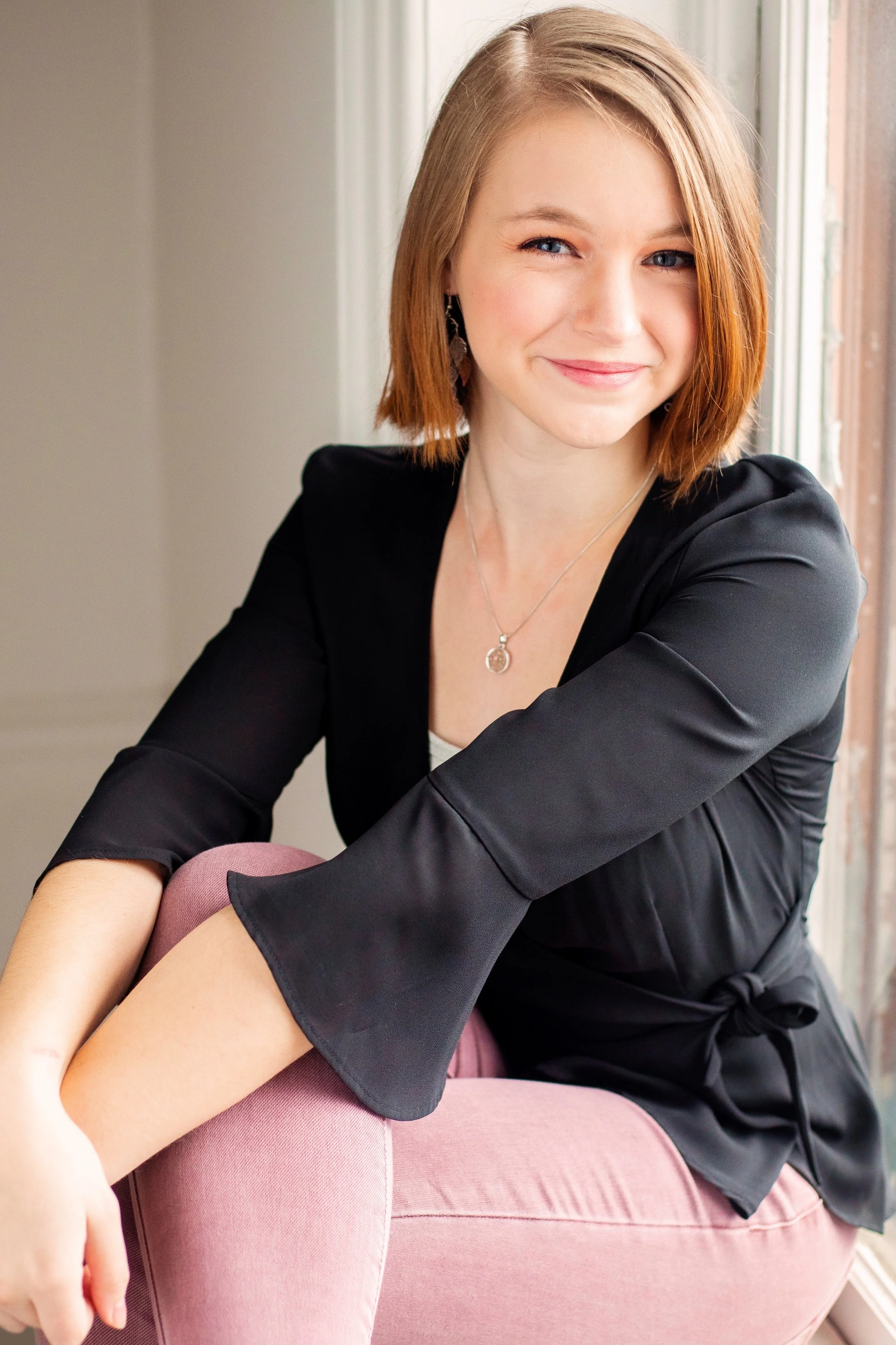 A woman with shoulder-length red hair sitting on a pink chair next to a window, wearing a black blazer, a necklace, earrings, and smiling at the camera.