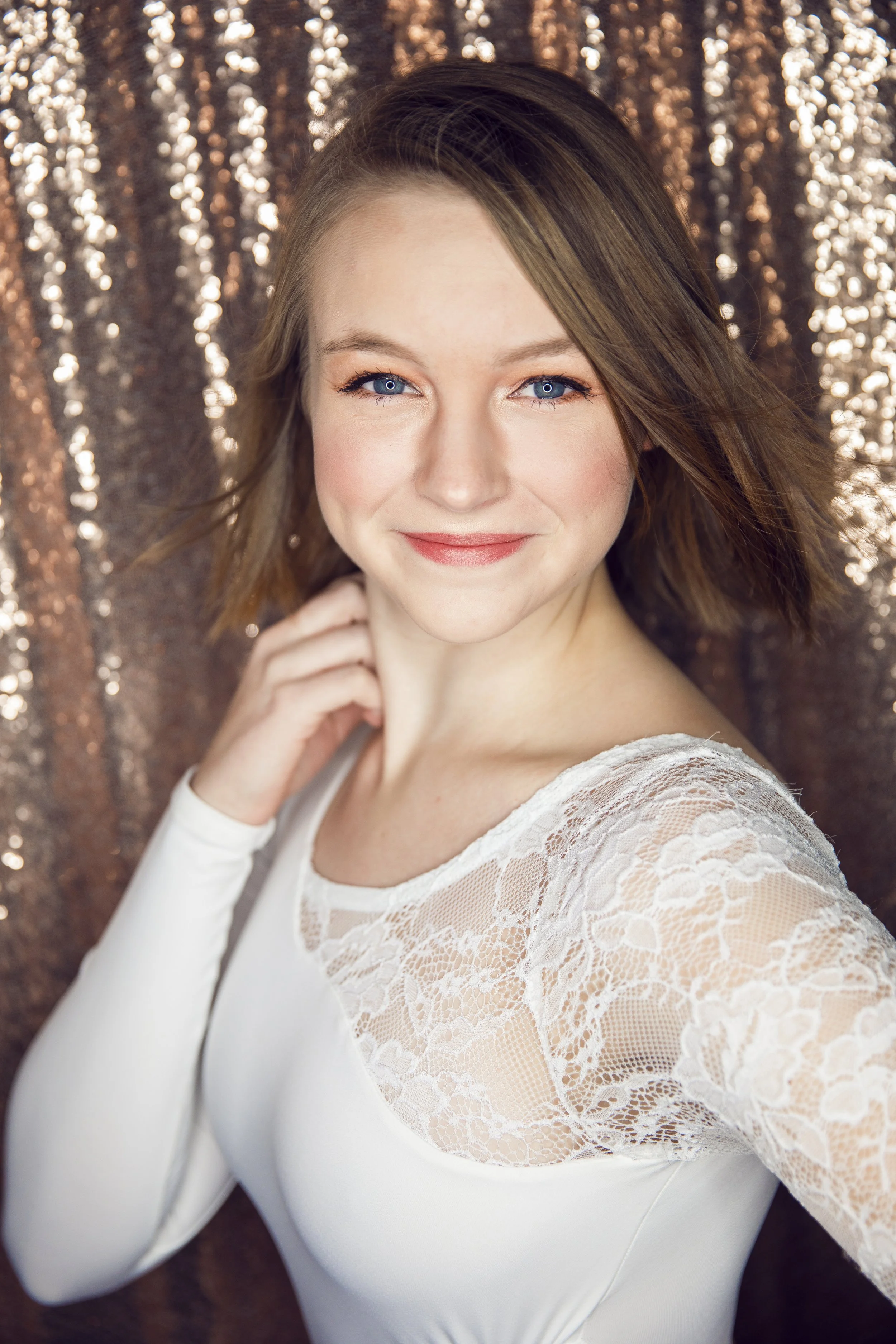 A woman with short, wavy brown hair, bright blue eyes, and a gentle smile, wearing a white dress with lace sleeves, standing in front of a sparkly, bronze-colored backdrop.