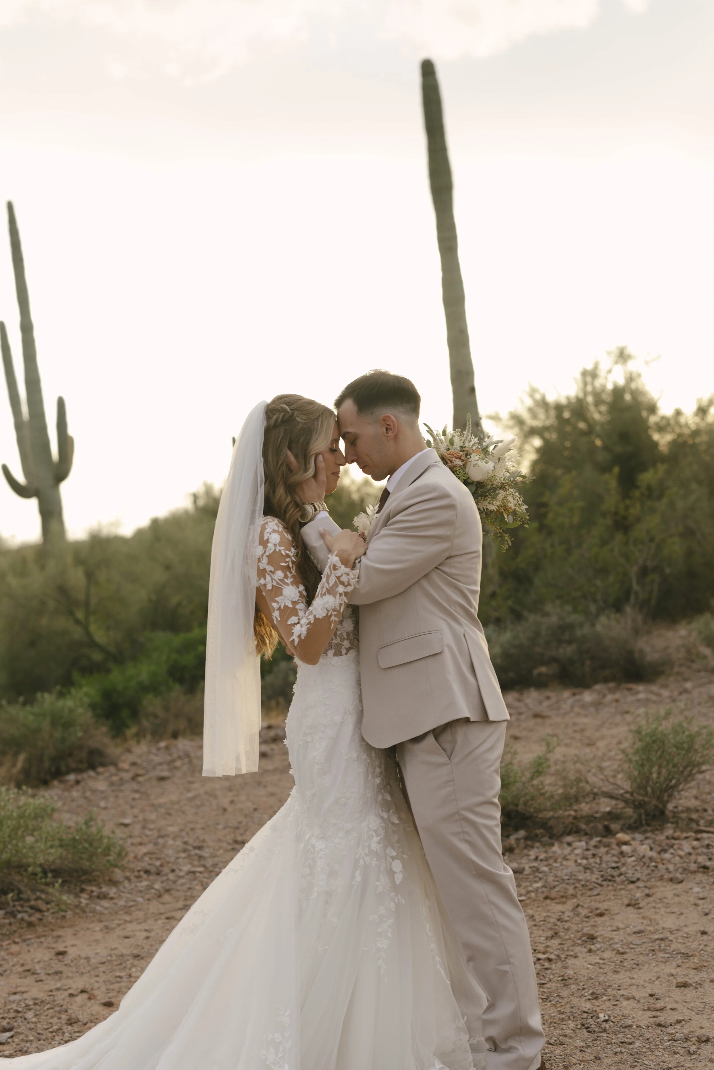 A bride and groom embrace in a desert landscape with tall cacti and greenery at sunset.