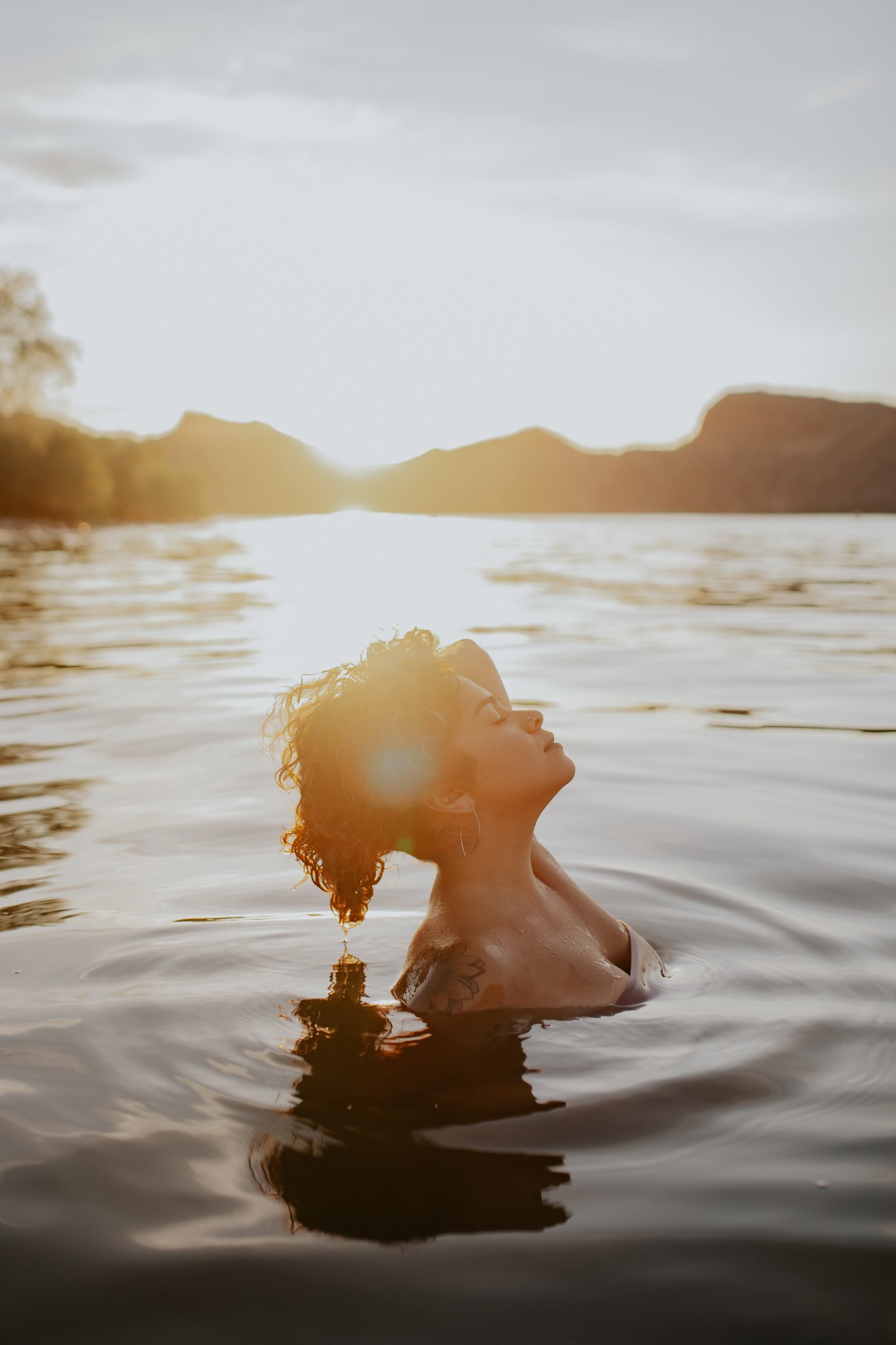 A woman with curly hair and earrings relaxing in a body of water during sunset, with mountains in the background.