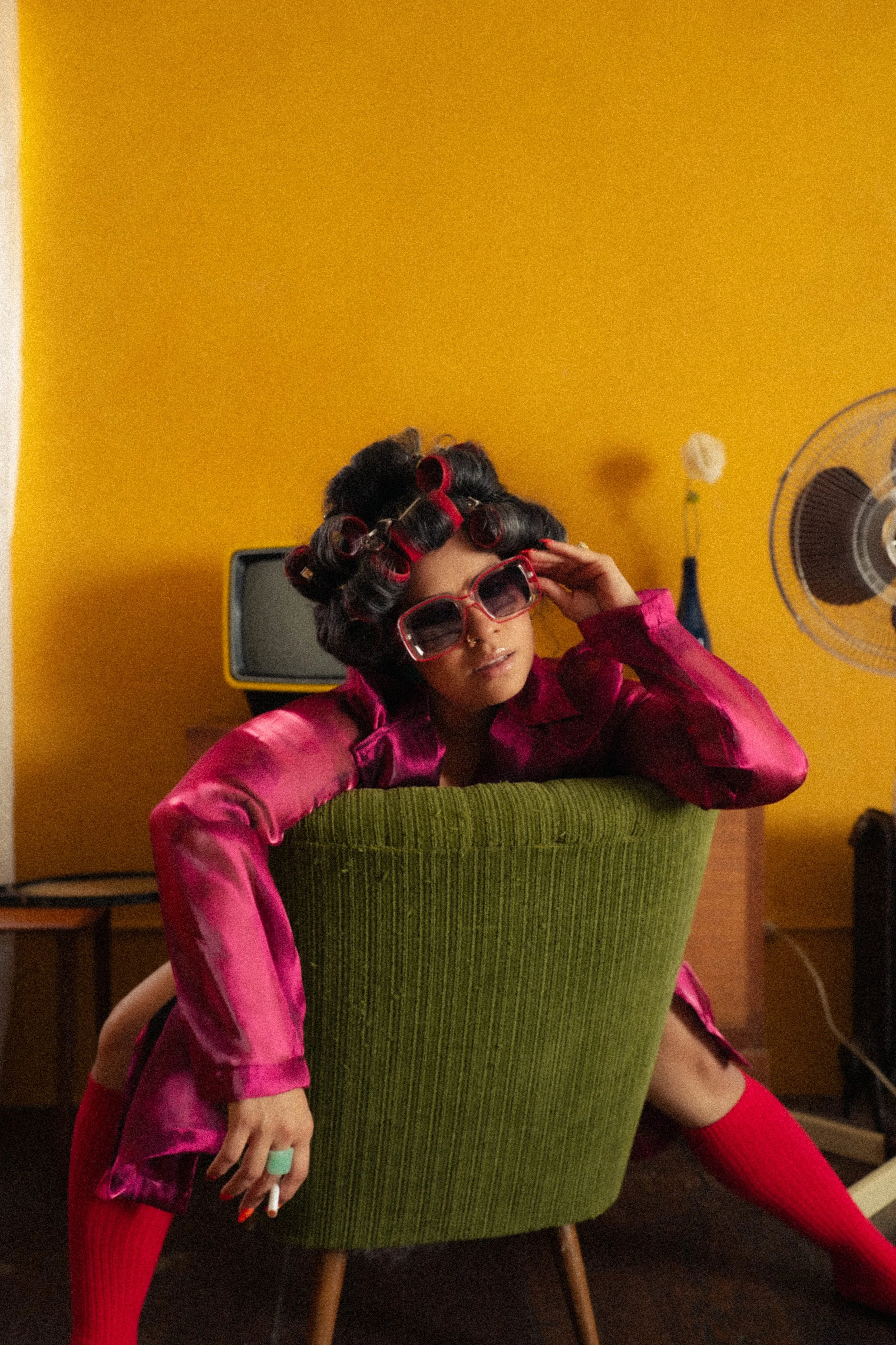 Woman with hair rollers and sunglasses sitting on a green chair in a retro-styled room with a yellow wall.