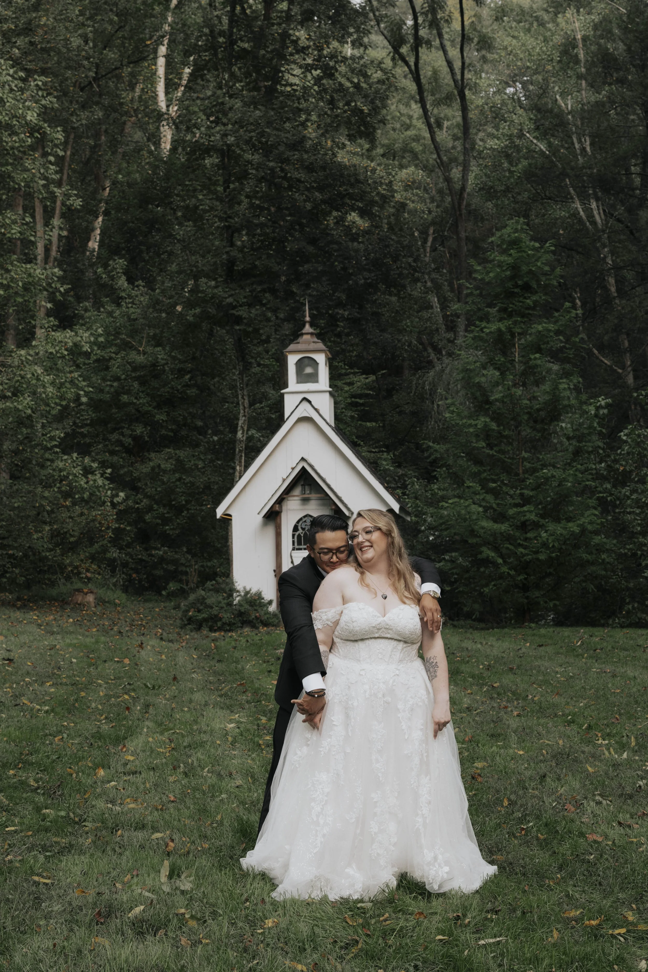 Two women in wedding gowns happily embracing outdoors in front of a small white chapel, surrounded by trees and greenery.