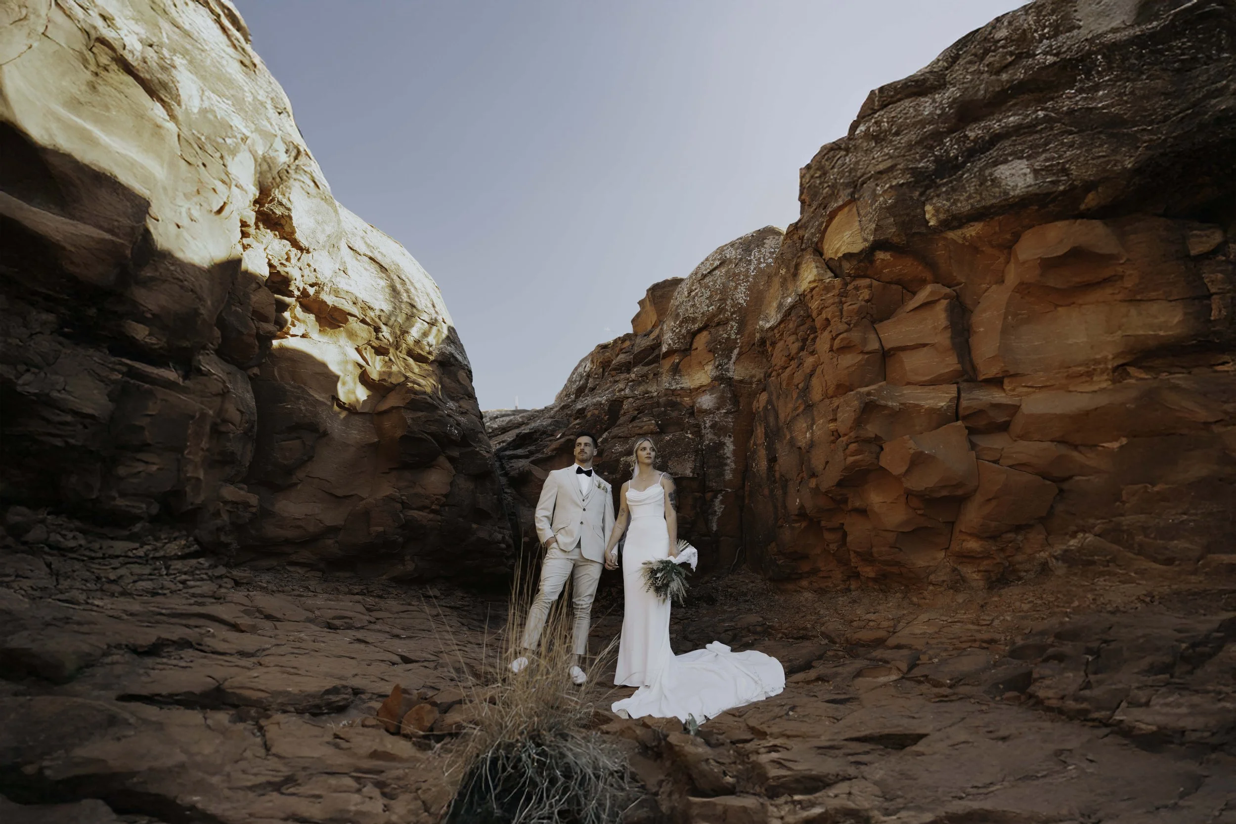 A bride and groom in wedding attire standing hand in hand in a rocky canyon, with the bride holding a bouquet and the groom in a beige suit with a black bow tie.