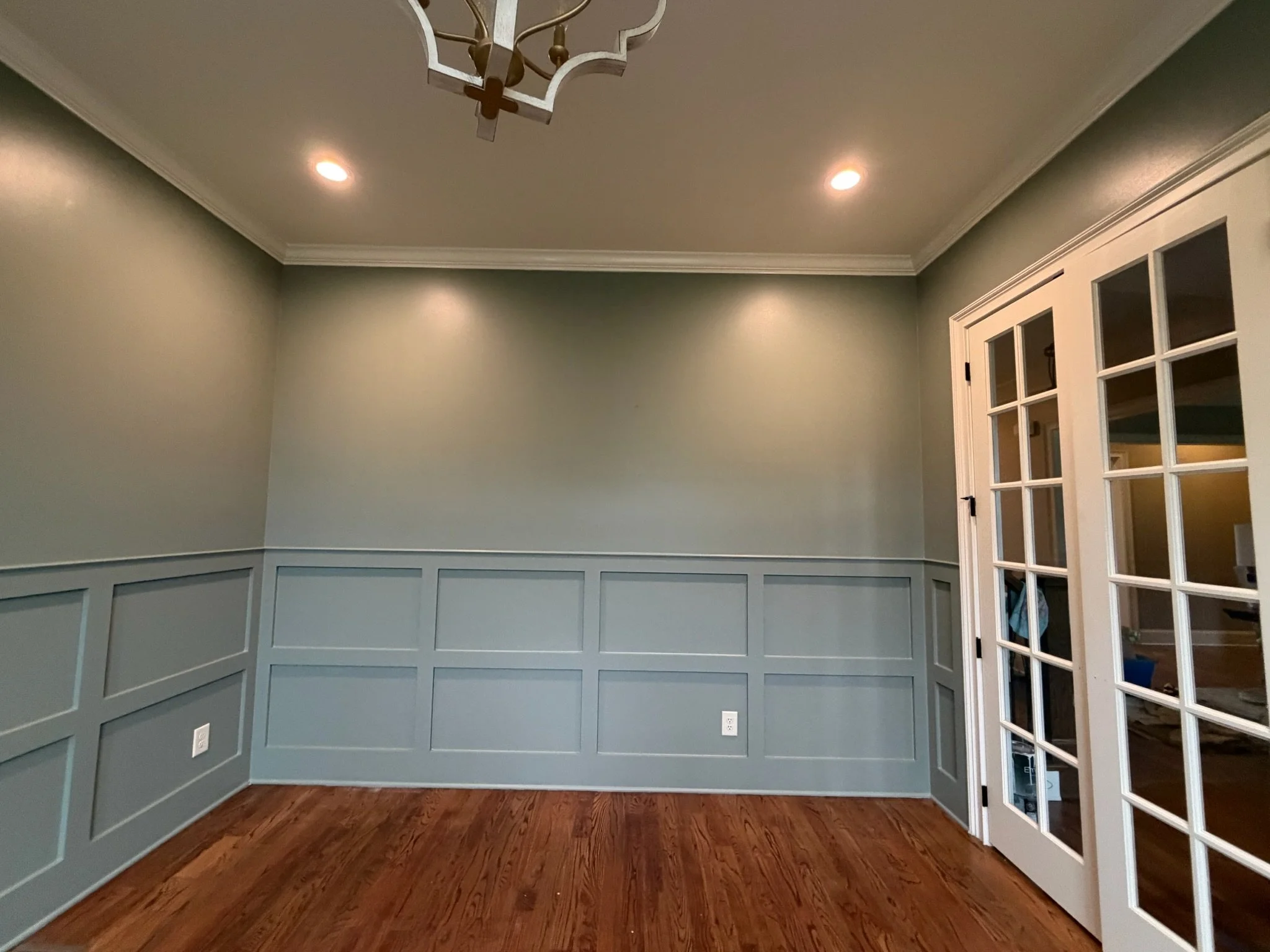 Empty dining room with hardwood floors, gray wainscoting, three ceiling lights, a chandelier, and a set of white French doors.