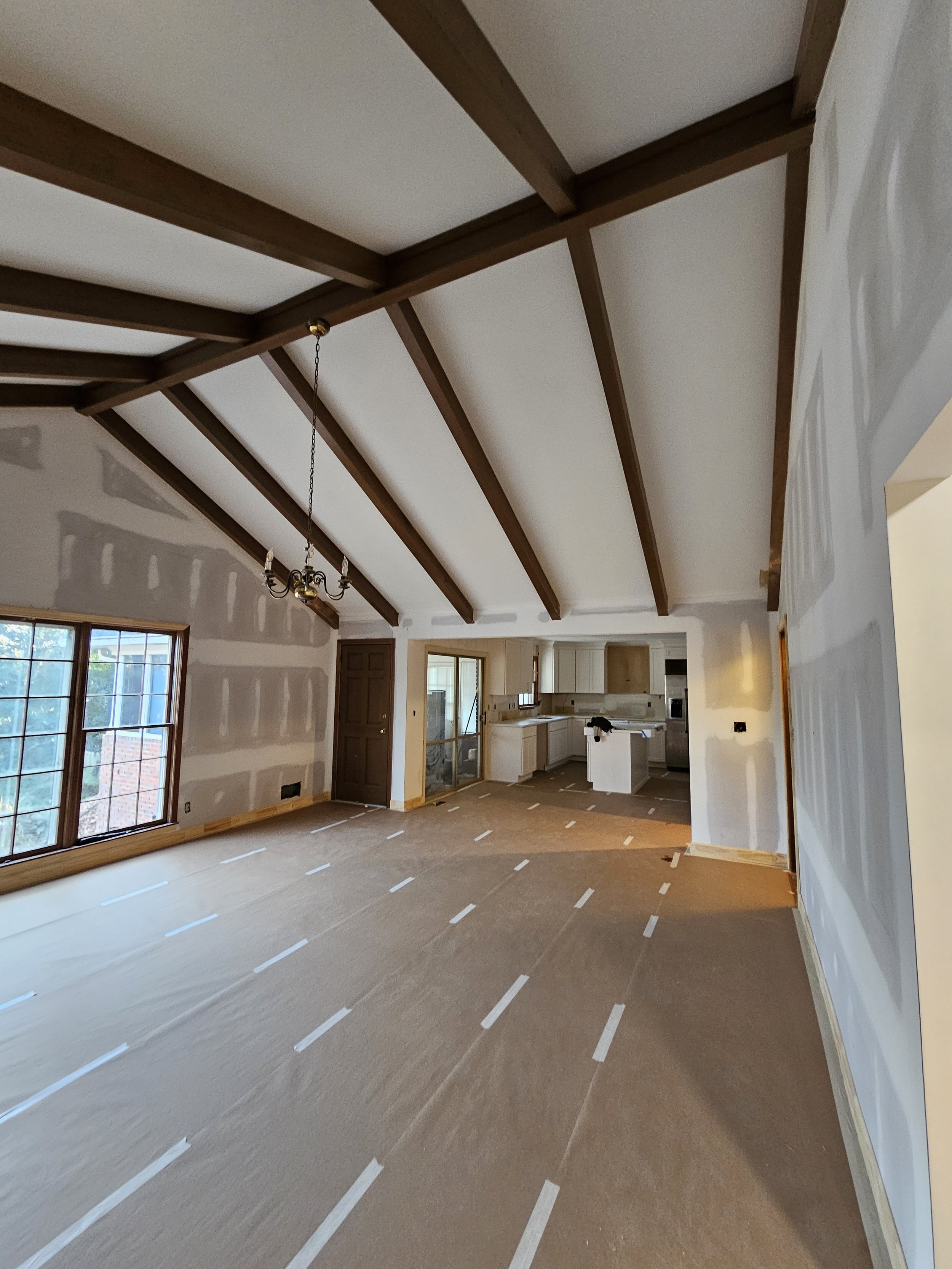 Interior view of a house under construction, showing drywall on the walls, exposed wooden beams on the ceiling, and a kitchen area in the background with white cabinetry and appliances. The floor is covered with protective paper marked with white tape.