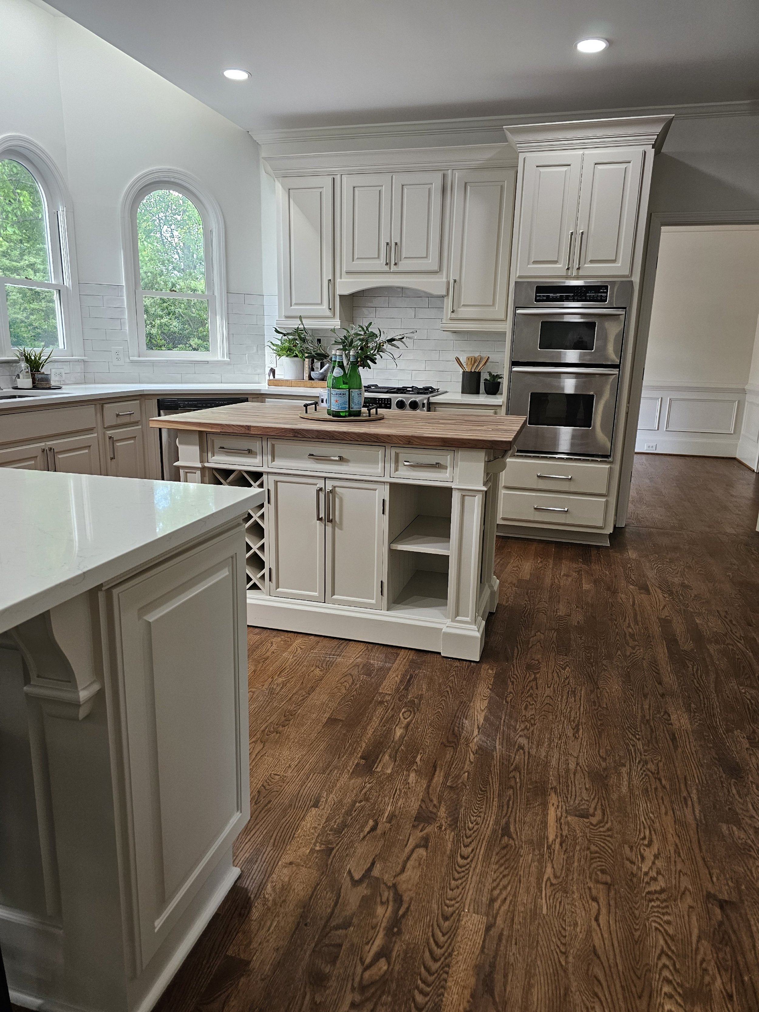 White kitchen with wooden flooring, white cabinetry, a kitchen island with a wooden top, and windows showing green trees outside.