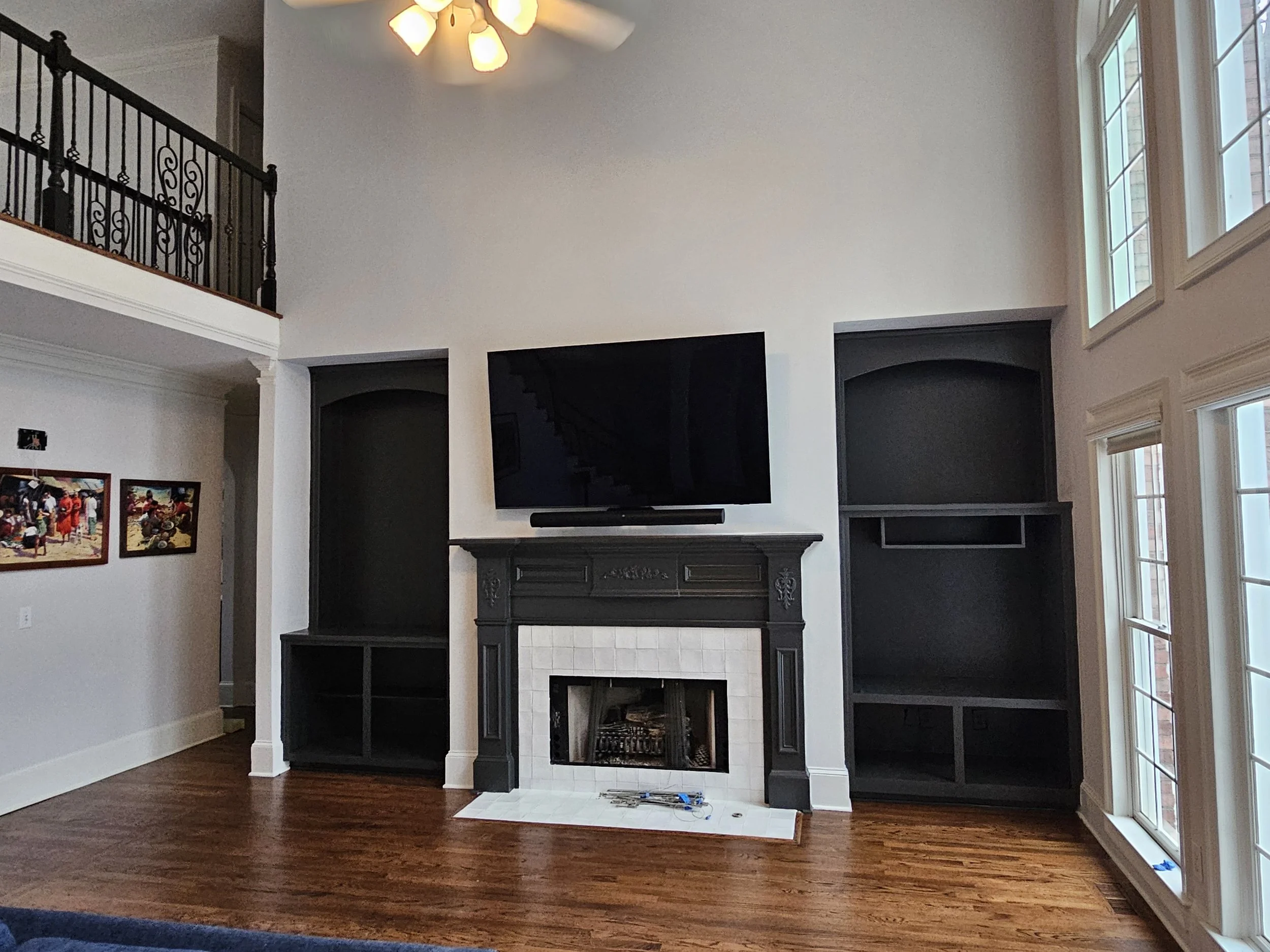Living room with a white wall, a black fireplace mantel, a wall-mounted flat-screen TV above the fireplace, dark built-in shelves on either side, large windows on the right, hardwood floor, and a ceiling light fixture.