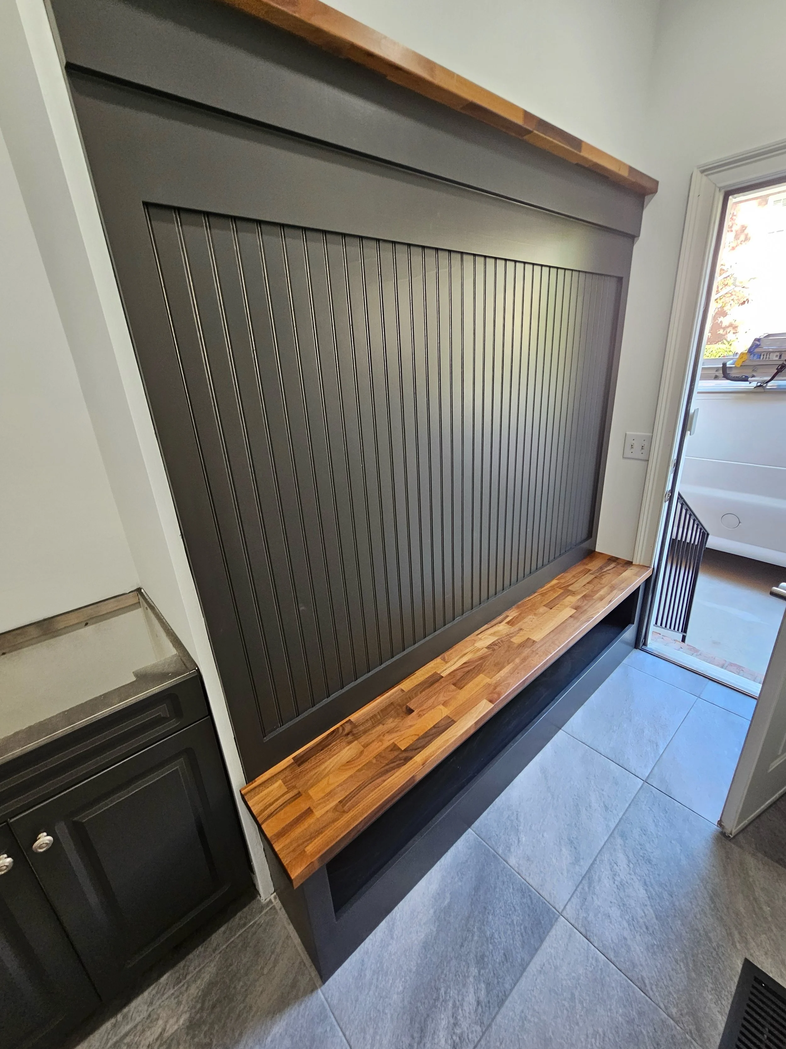 A modern interior with a black beadboard feature wall, a wooden shelf, and a small black cabinet. An open door reveals a bathroom with a white bathtub, and beside it is a patio with a railing.