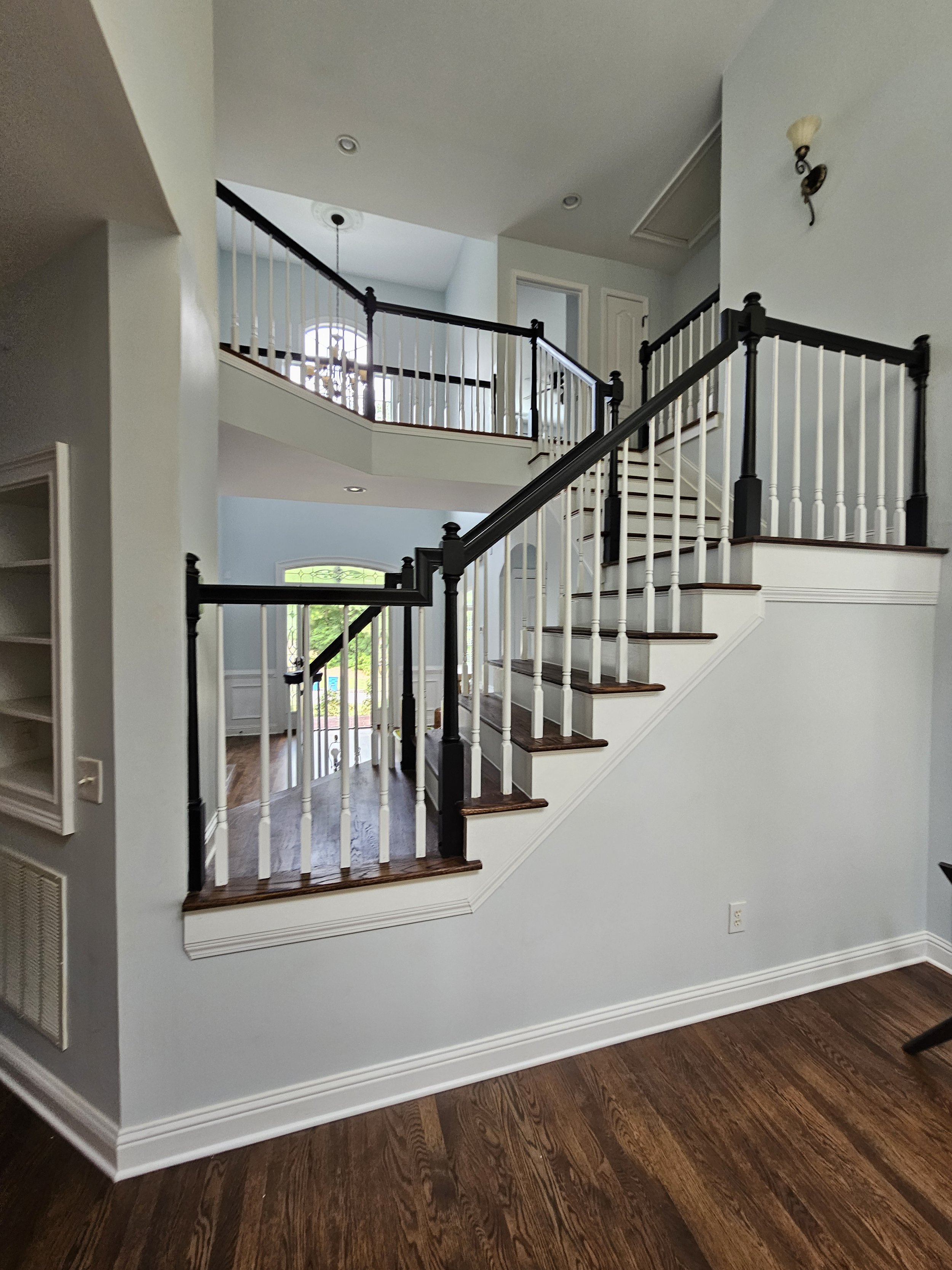 Interior of a house showing a staircase with black and white railing leading to an upper floor, hardwood floors, a wall with built-in shelves, and a doorway with a window letting in natural light.