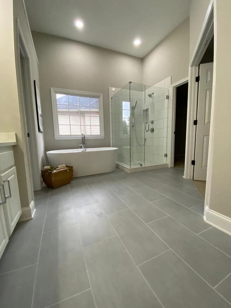 A modern bathroom featuring a freestanding bathtub, a glass-enclosed shower, a window above the bathtub, and light-colored tiled flooring.
