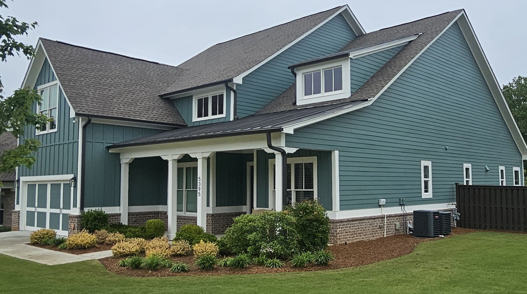 A two-story house with teal siding, white trim, and a gray shingle roof, surrounded by a manicured lawn and landscaped bushes.