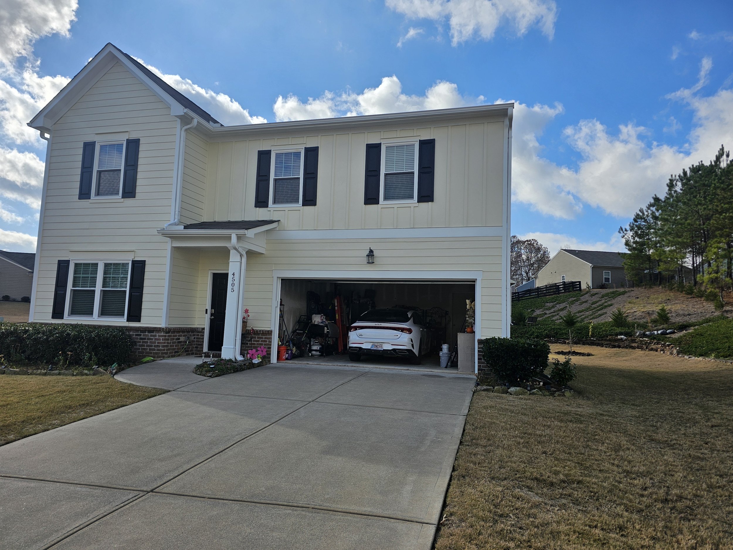 Front view of a two-story yellow house with white trim, black shutters, and a partially open garage door with a white sports car inside. The house is on a paved driveway, with a lawn and landscaping on the sides, and a blue sky with clouds overhead.