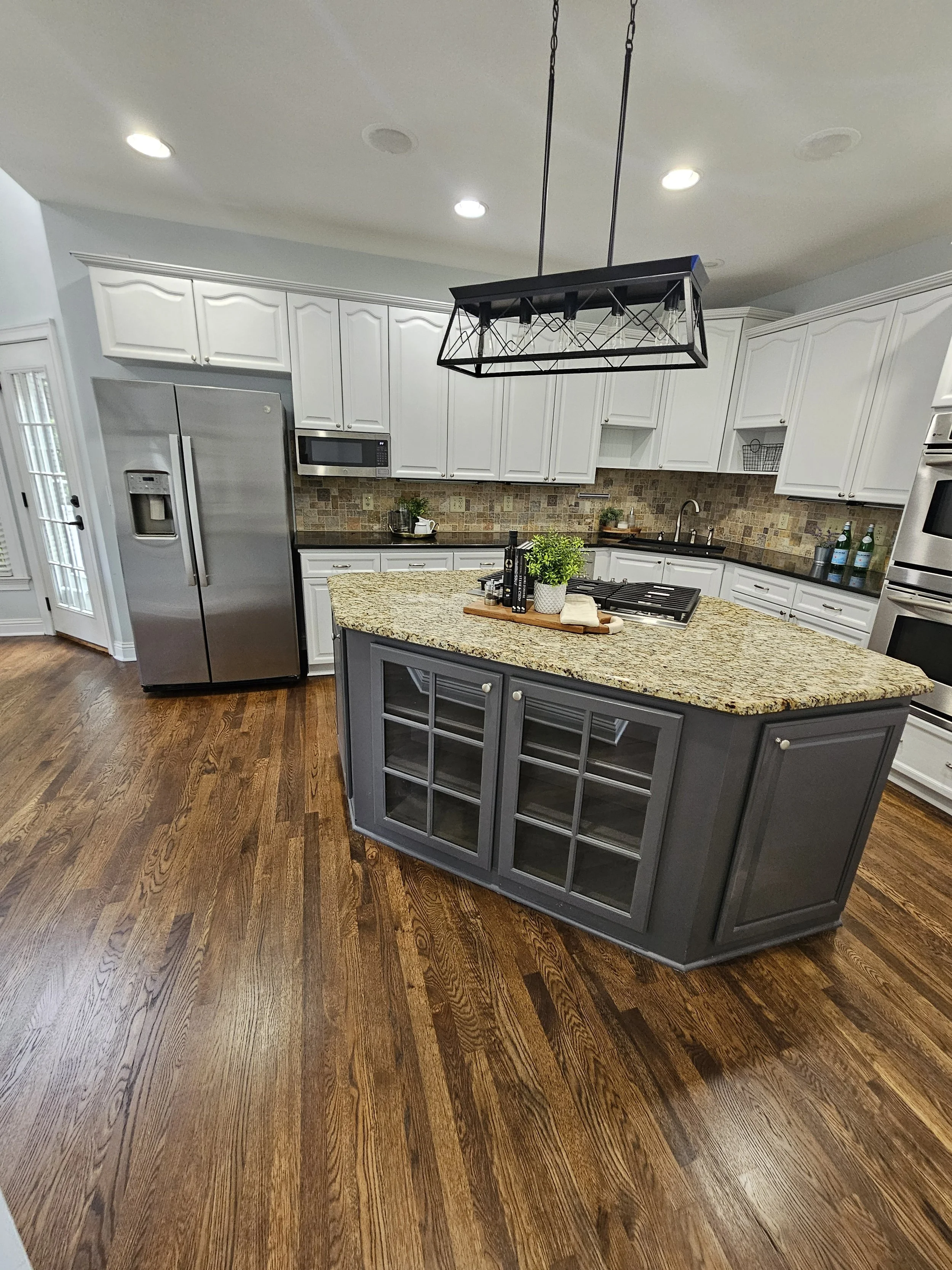 A modern kitchen with white cabinets, stainless steel refrigerator, granite island with gray base, and hardwood floors.