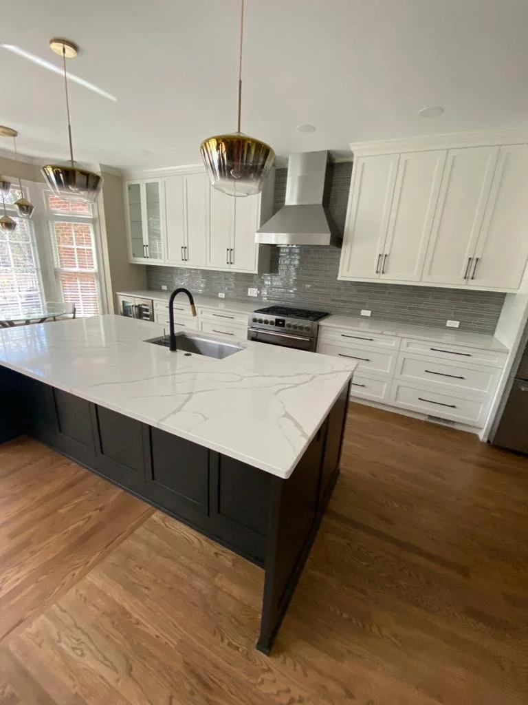 Modern kitchen with white cabinets, a black island countertop with a white marble pattern, stainless steel appliances, gray tiled backsplash, wooden floors, and gold pendant lights.