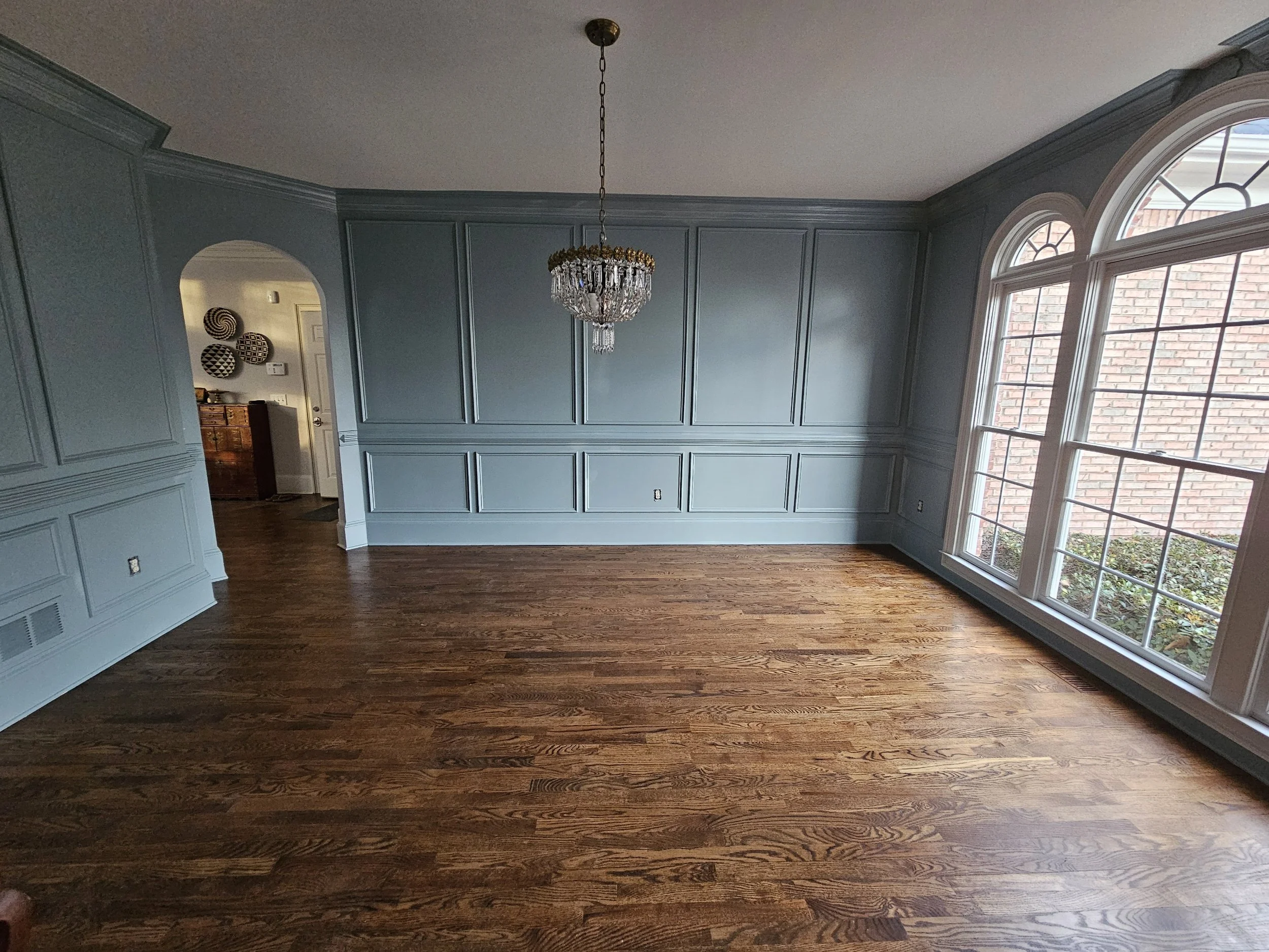 Empty living room with large front windows, blue painted panel walls, and a chandelier hanging from the ceiling.