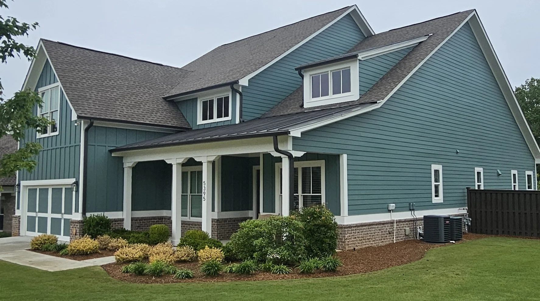 A two-story house painted in blue with white trim, a small front porch, manicured lawn, flower beds, and an HVAC unit in the backyard.