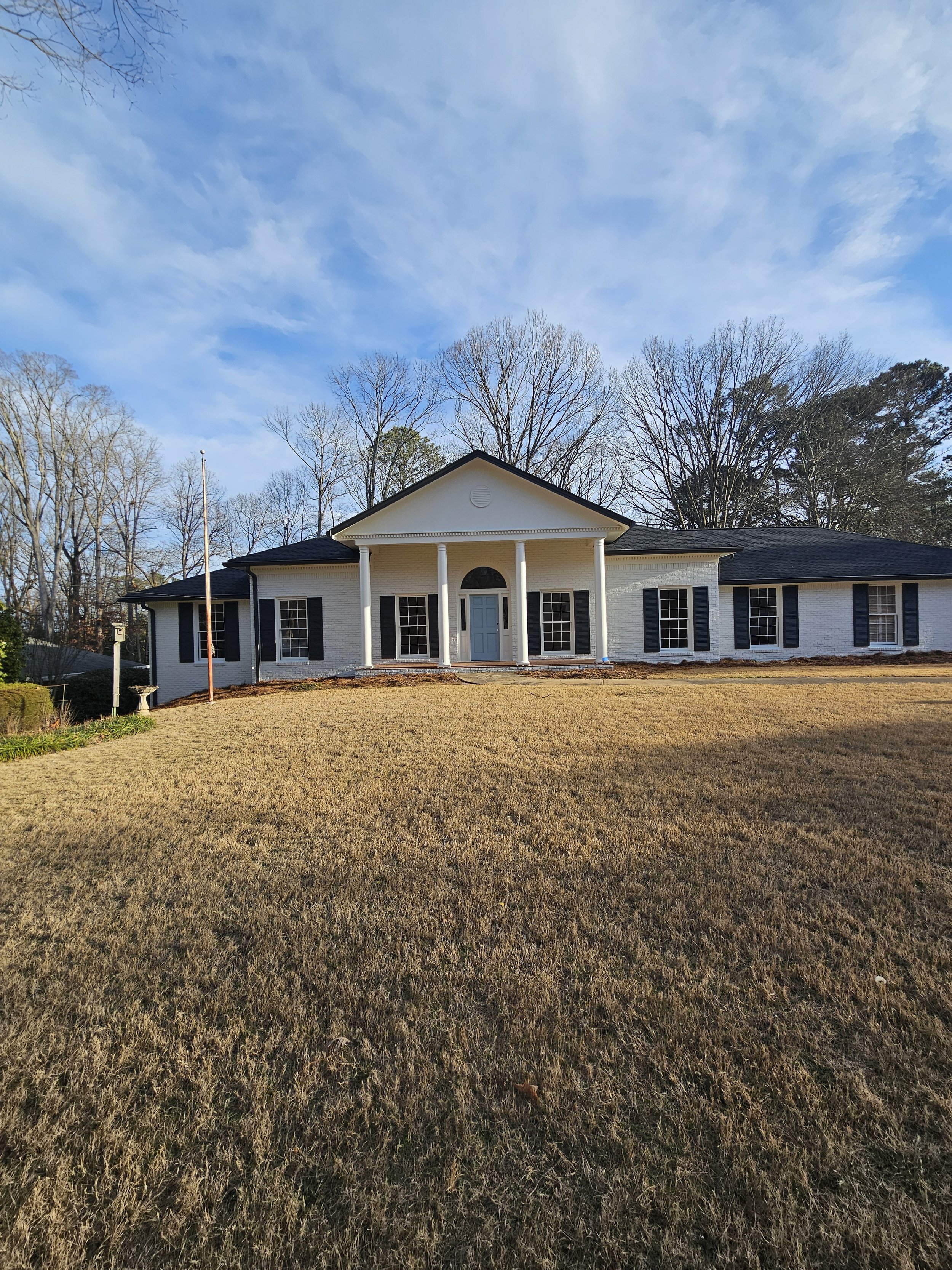 Front view of a white brick house with black shutters, a central front porch with columns, and a grass lawn in the foreground. Trees with bare branches are behind the house, and a blue sky with scattered clouds is overhead.