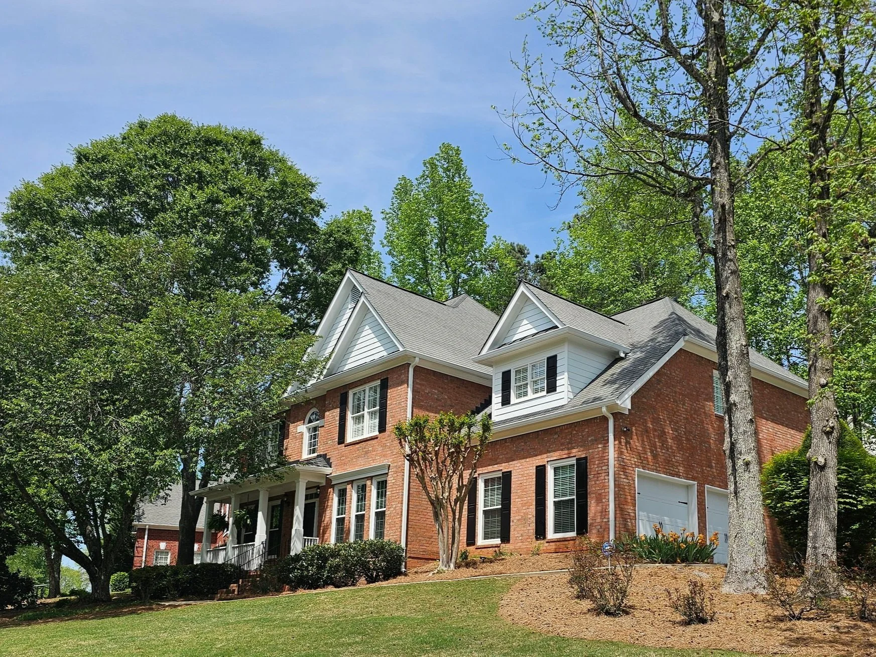 Large brick house with multiple roof peaks, white trim, black shutters, and a front porch surrounded by trees and greenery under a blue sky.