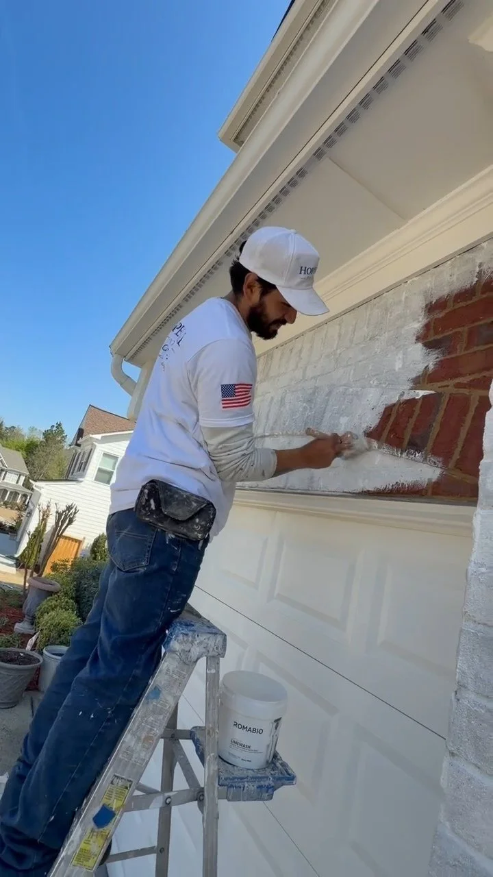 A man painting the exterior brick wall of a house on a ladder during daytime, wearing a white cap and t-shirt, with a bucket of paint nearby.