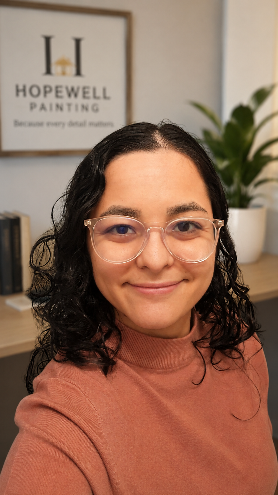 A woman with dark curly hair and glasses smiling, wearing a rust-colored top, with a framed artwork and a plant in the background.