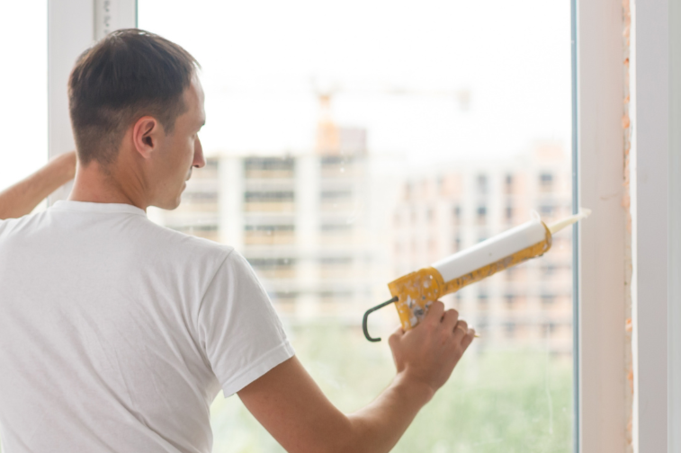 A man in a white shirt applying white sealant or caulk to a window frame using a caulk gun.