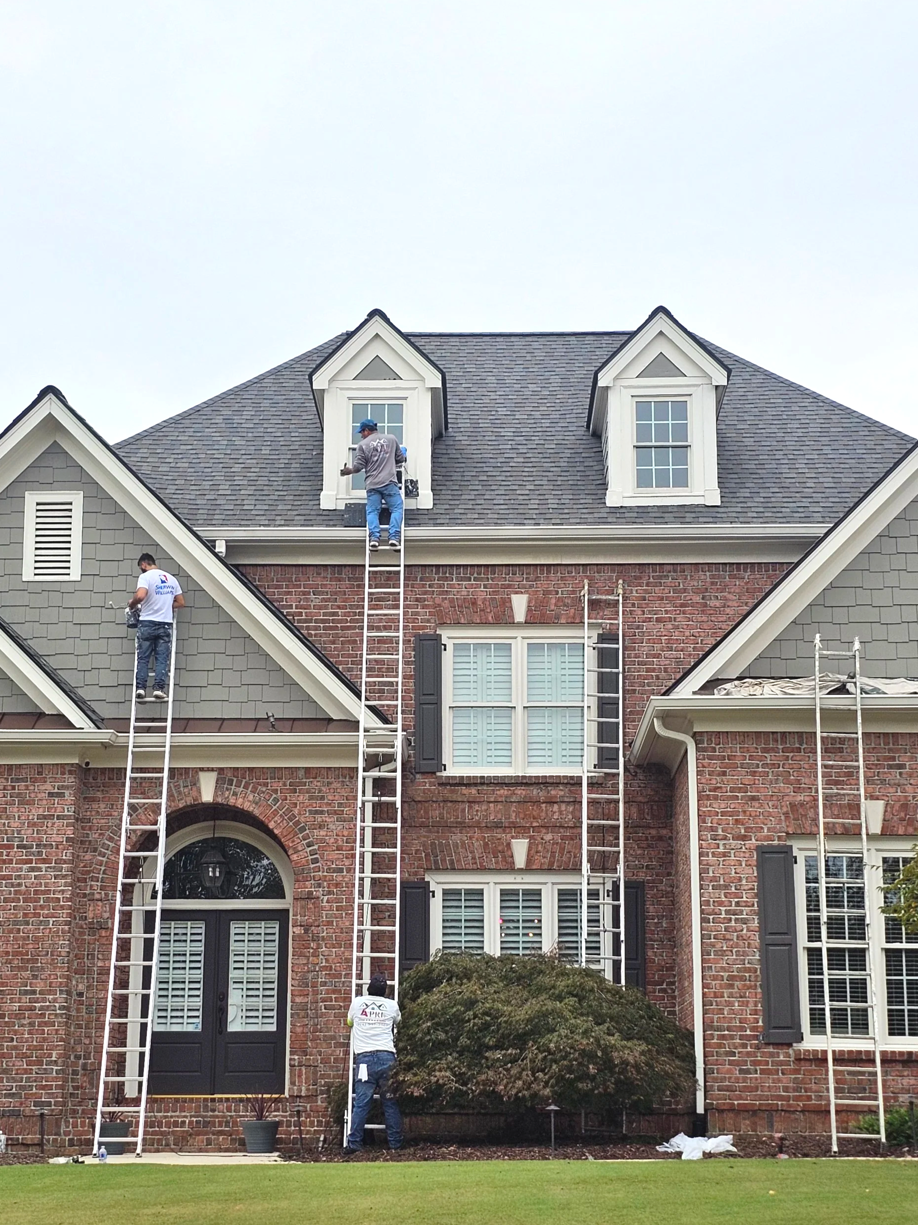 Workers installing or repairing roof on a brick house using ladders and scaffolding, with one worker on the roof and others below.