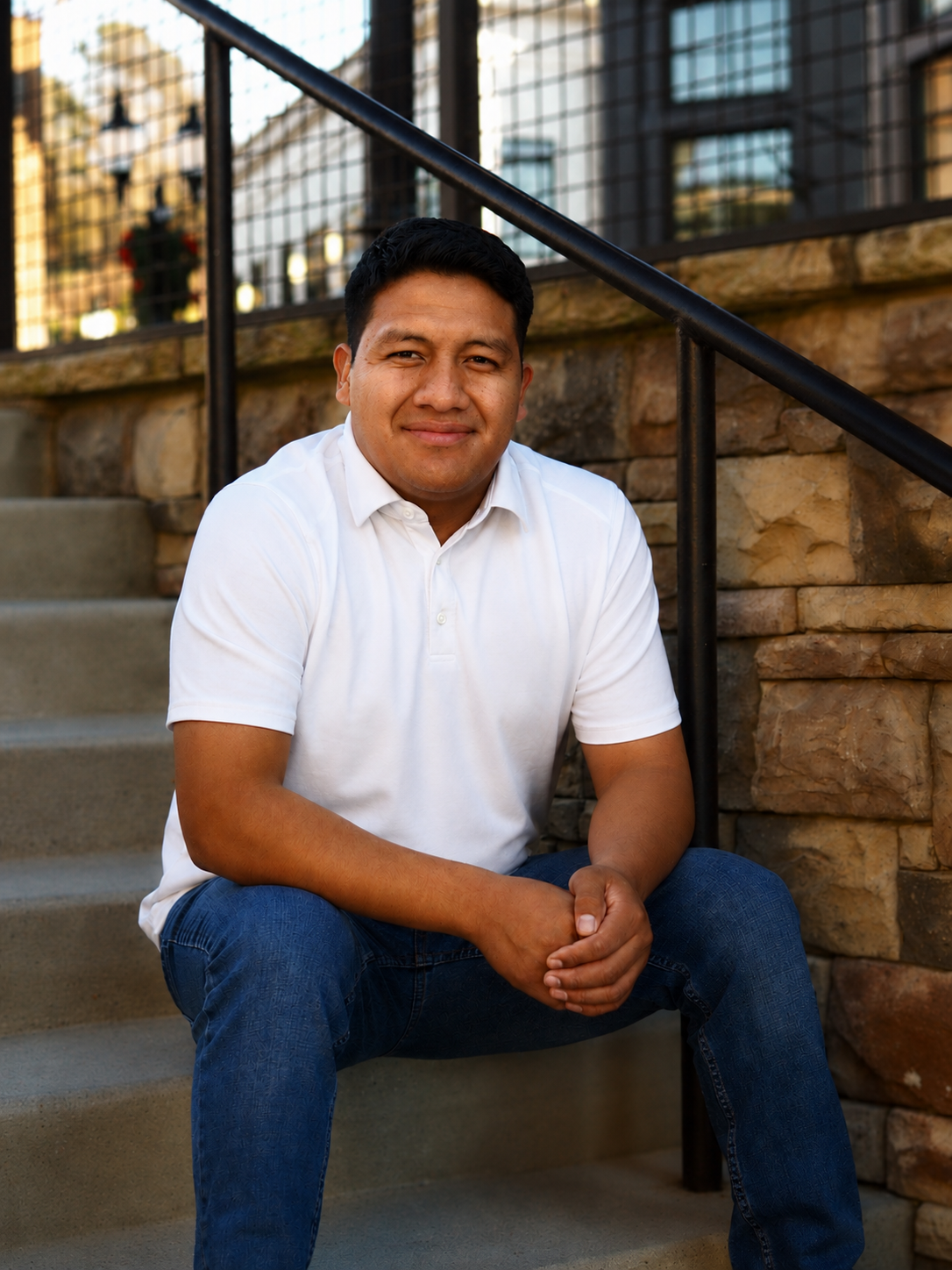 A man sitting on outdoor concrete stairs, wearing a white polo shirt and blue jeans, smiling at the camera, with a black metal railing and a brick wall behind him.