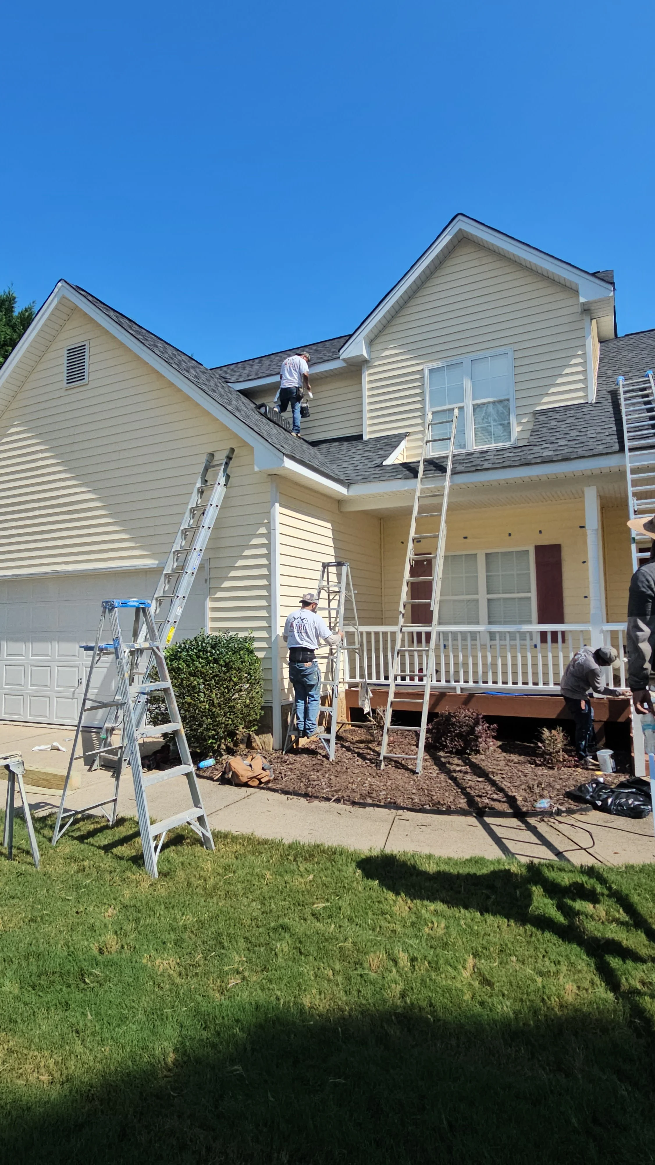 Workers repairing the roof of a yellow two-story house with ladders and tools under a clear blue sky.