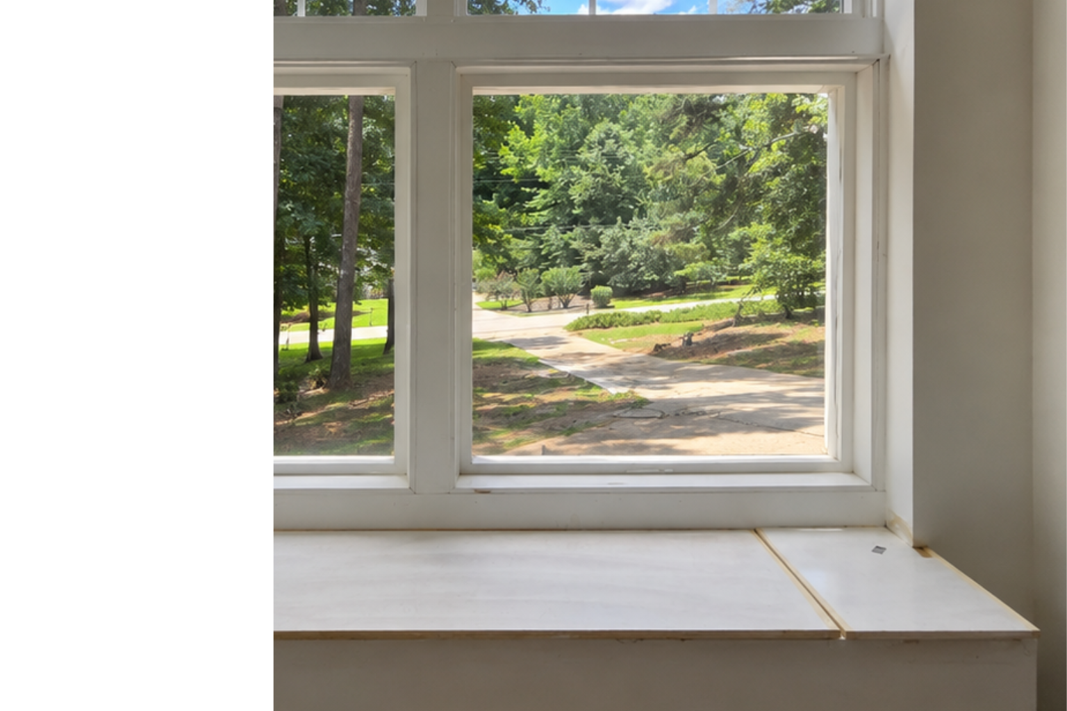 View of a sunny suburban neighborhood with trees, houses, and a sidewalk through a window with a white sill.