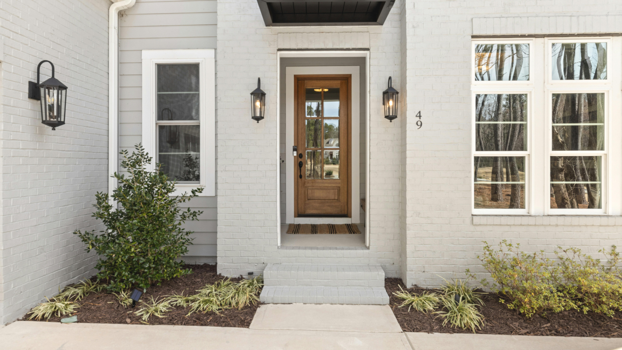 Front entrance of a modern house with a wooden door, brick and siding exterior, black wall-mounted lanterns, window, number 49, steps leading up to door, landscaped yard with bushes and plants.