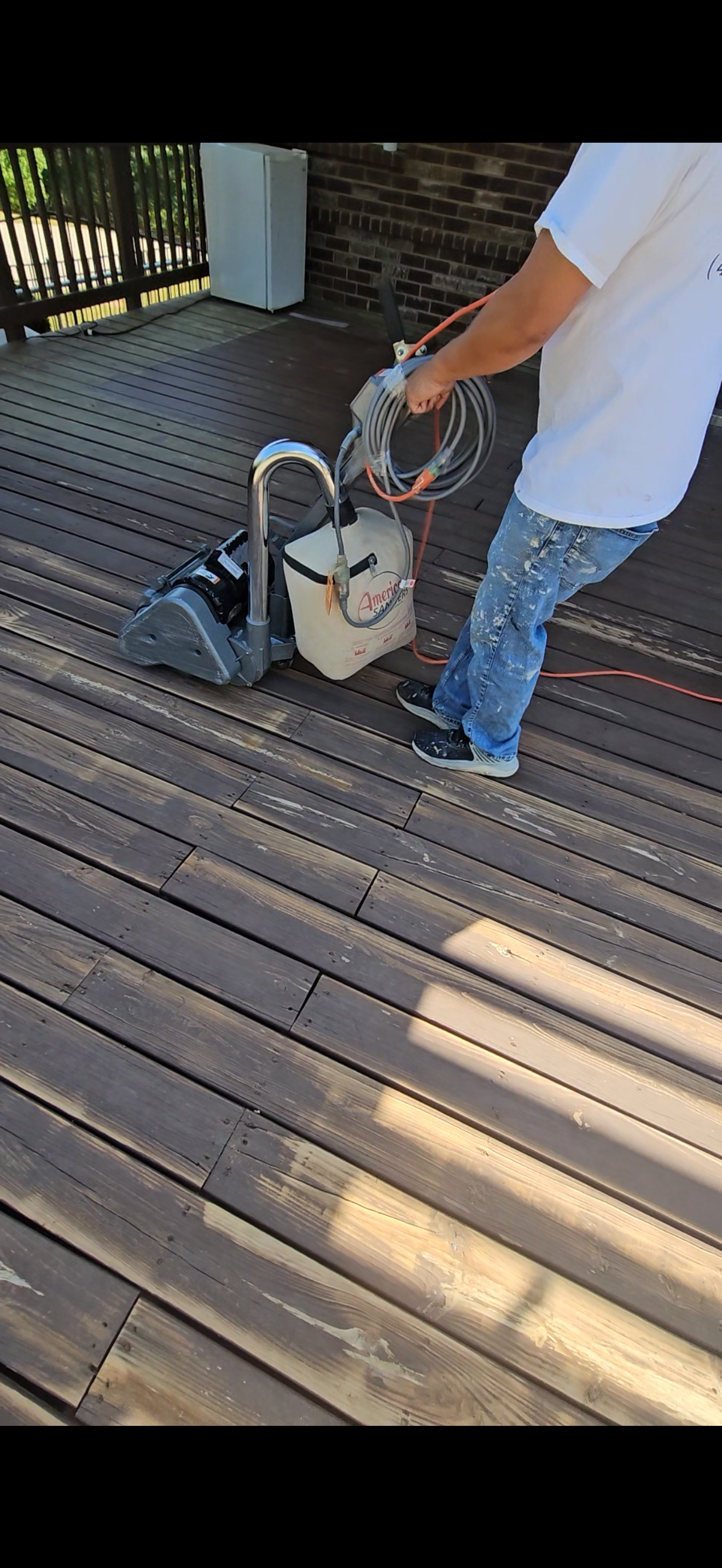 A person working on wooden deck flooring using a powered sander, wearing a white t-shirt and blue paint-splattered jeans.