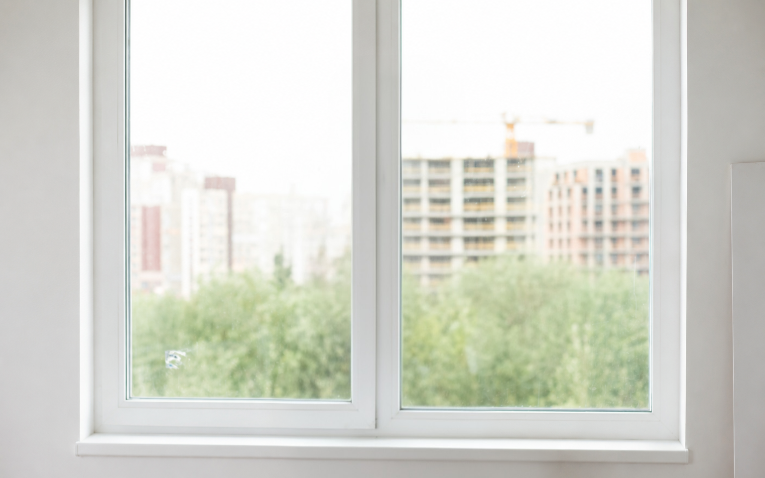 View of a cityscape through a glass window with white frame, showing buildings and green trees outside.