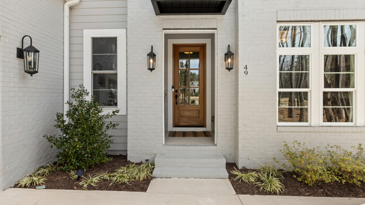 White brick house entrance with wooden door, glass panes, and outdoor lanterns, numbered 49, with plants and shrubs on either side.