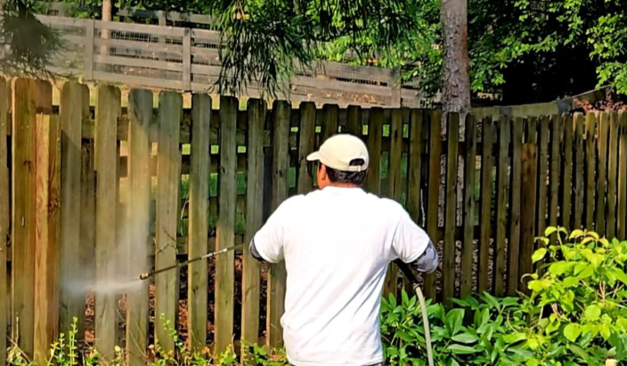 A person wearing a white T-shirt and white cap is pressure washing a wooden fence in a backyard with green trees and plants.