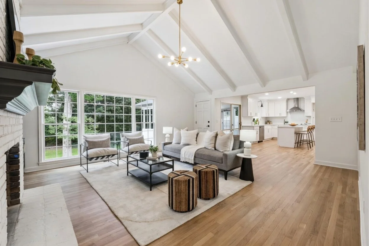 Bright living room with large window, white walls, vaulted ceiling with exposed beams, in a modern home. Contains a grey sofa with multiple throw pillows, a coffee table, two striped a ottomans, two armchairs, and side lamps. Open kitchen with white cabinetry and bar stools visible in the background.