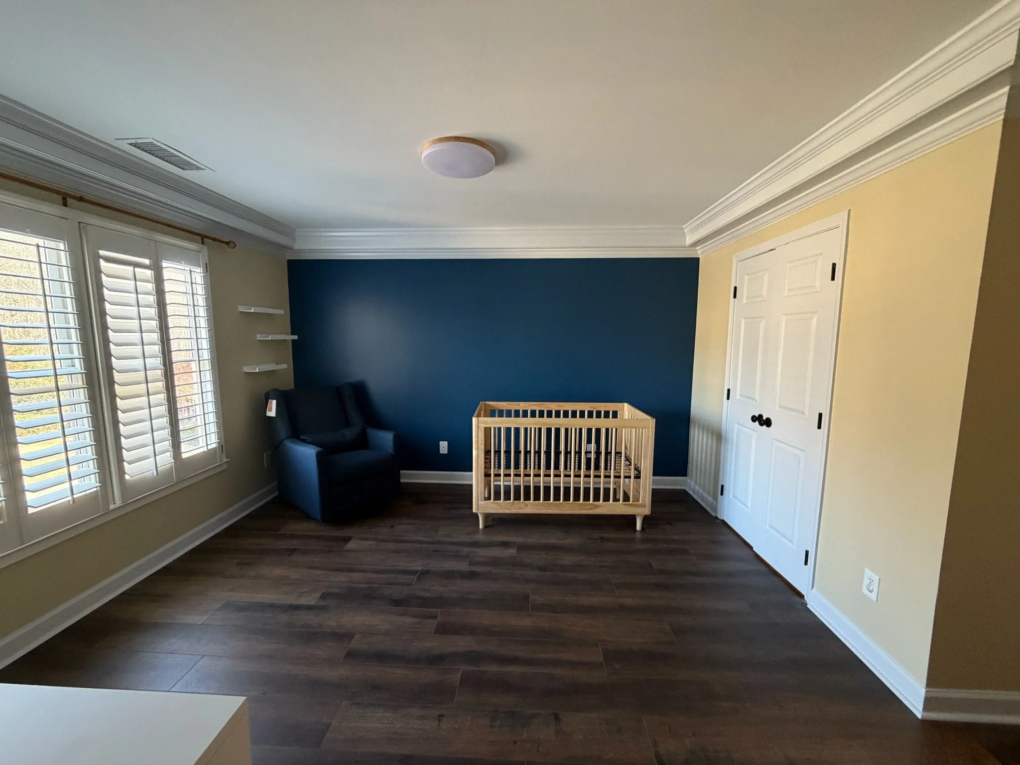 Empty nursery room with a blue accent wall, a wooden crib, a dark armchair, bookshelf shelves, windows with shutters, and a closet door.