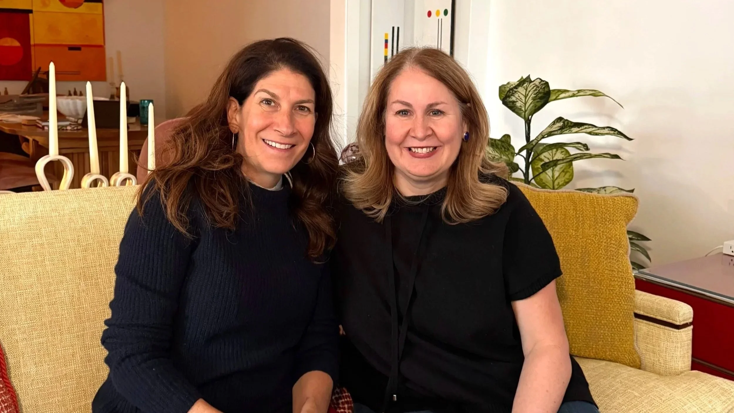 Two women sitting close together on a yellow sofa, smiling at the camera, with a houseplant and candles in the background.