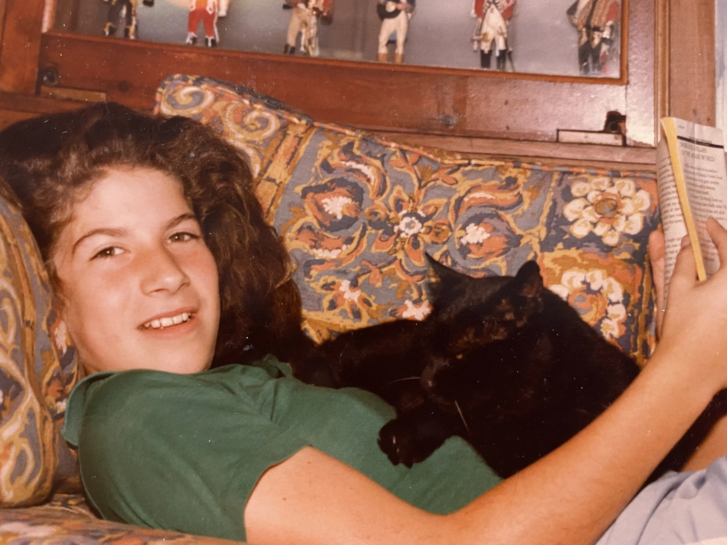 A girl with curly brown hair lying on a floral patterned couch, reading a book, with a black cat resting on her arm and side.
