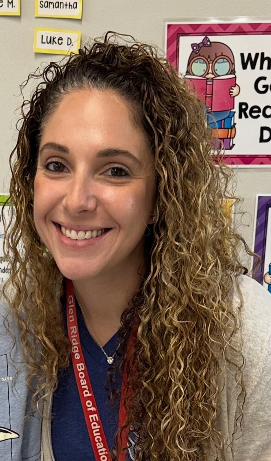 A woman with curly hair smiling in front of a classroom wall with educational posters and labels.