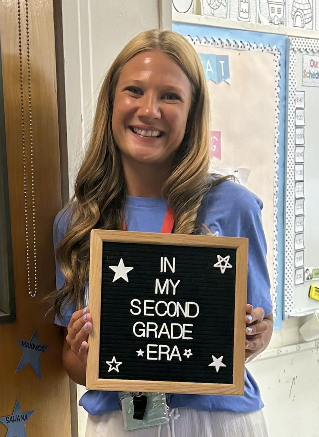 A smiling woman with long blonde hair holding a black letter board that says 'In My Second Grade Era' with star and heart designs, standing in a classroom.
