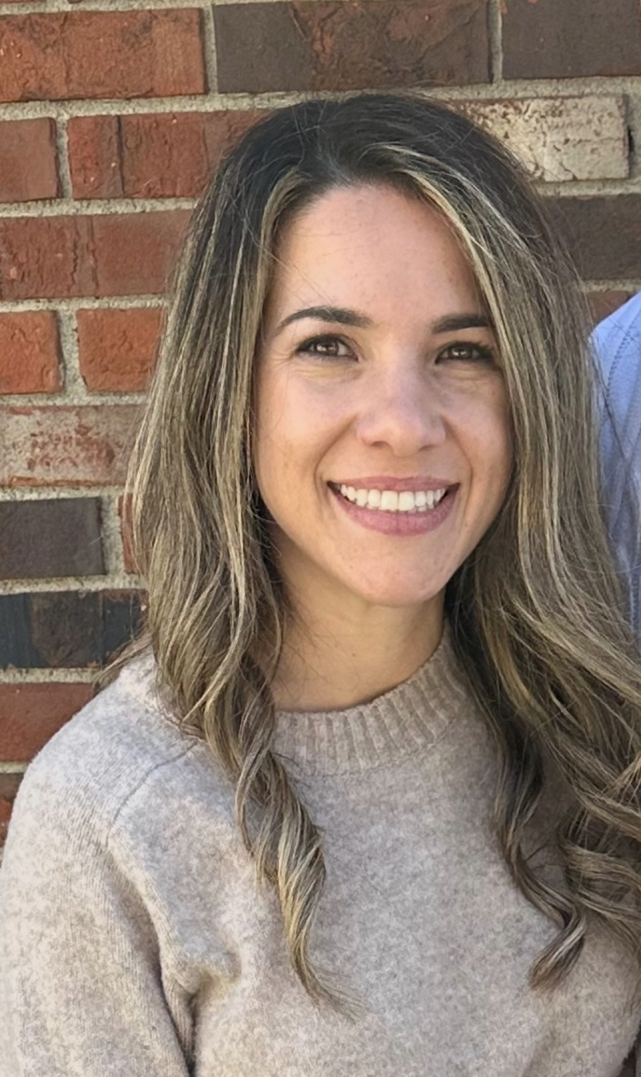 A woman with wavy, shoulder-length hair smiling in front of a brick wall.