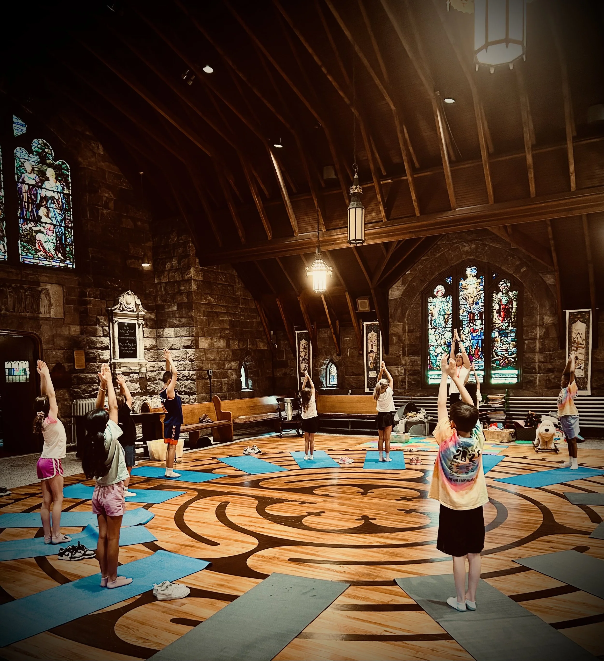 Children participating in a yoga class inside a church with stained glass windows and wooden ceiling beams, standing on individual mats, raising hands above heads.
