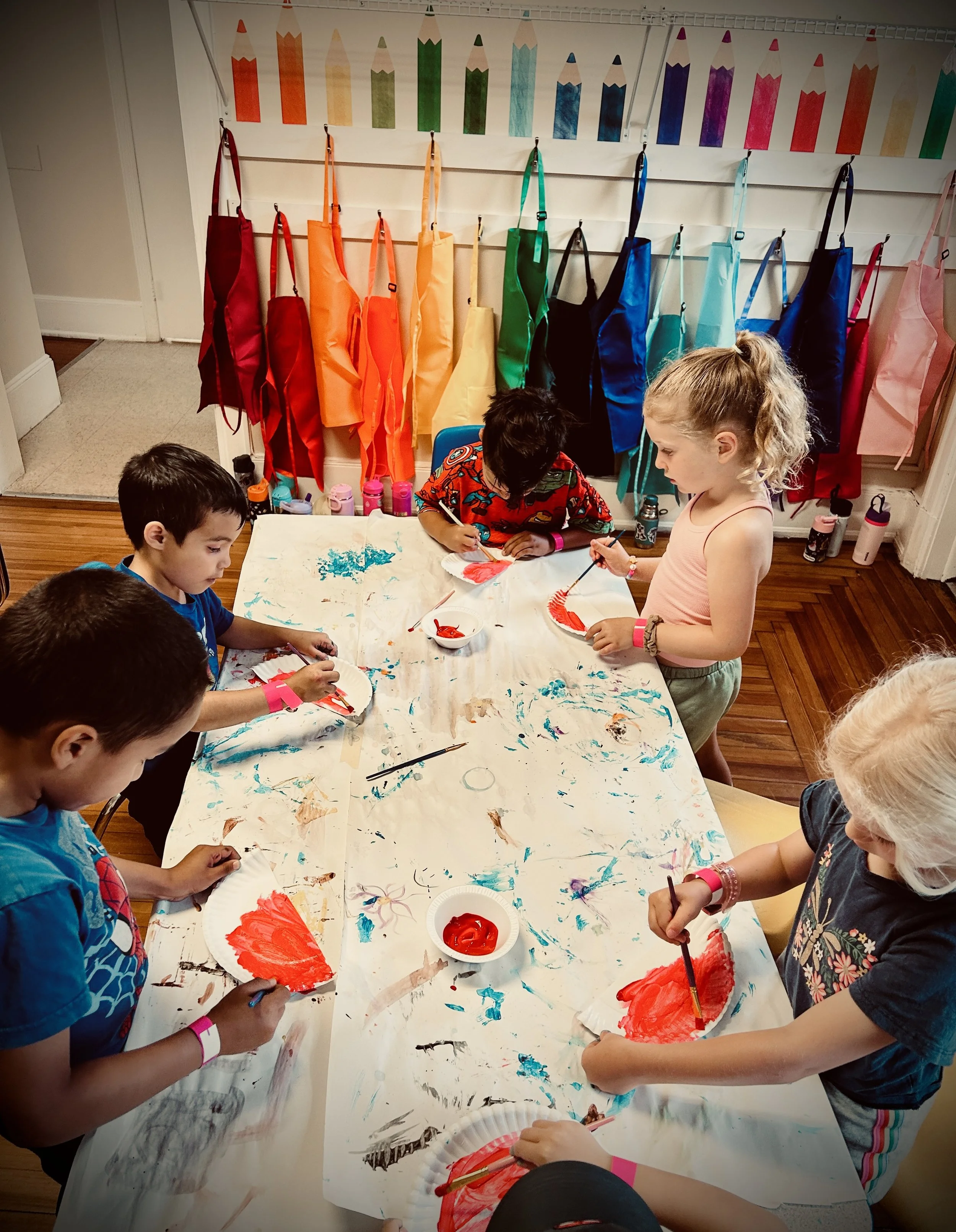 Children painting and drawing on paper with red and blue paint at a table, with colorful pencil-shaped decorations hanging on the wall behind them.