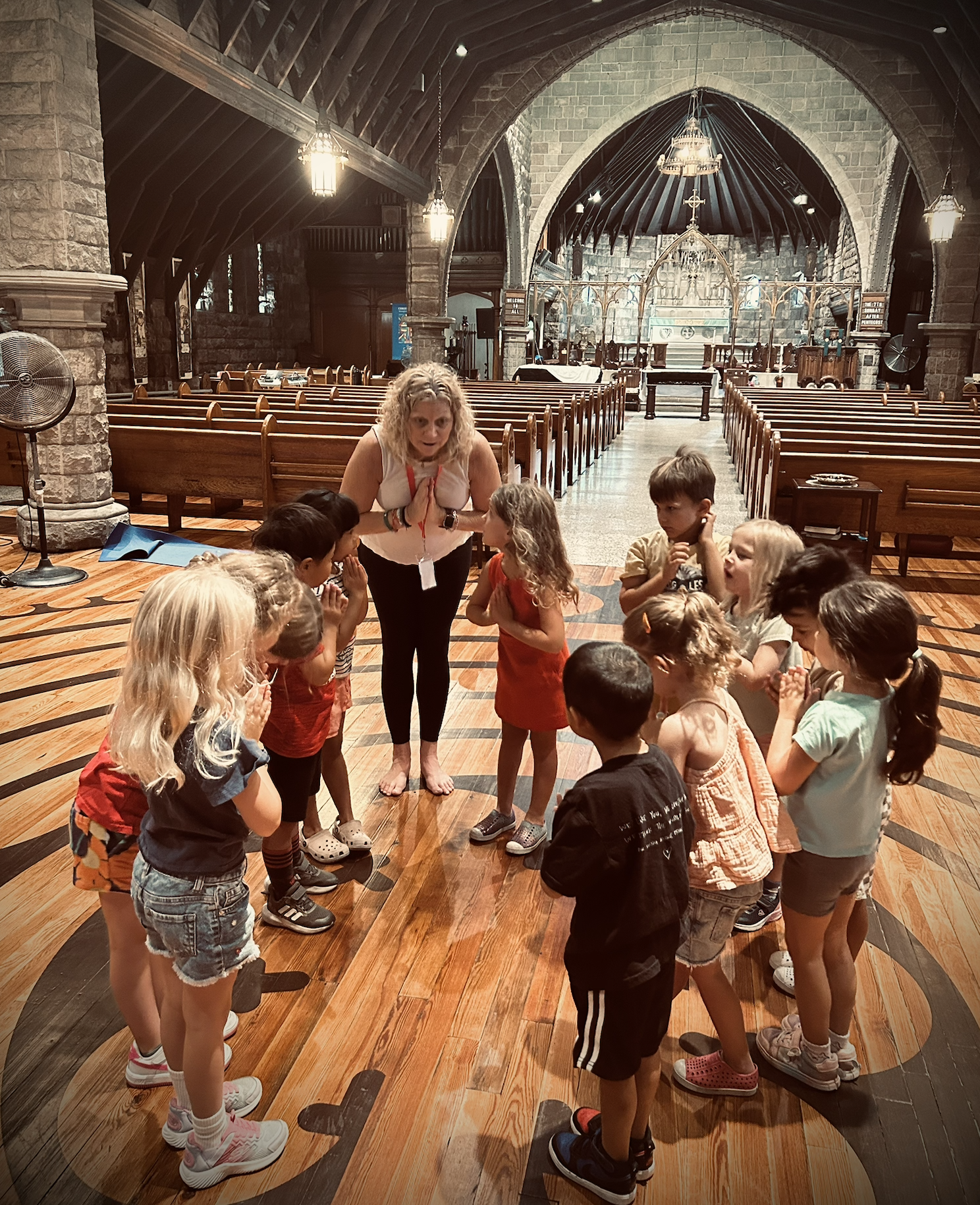 A group of children gathered in a circle around a woman inside a church with wooden pews and stone walls, with an altar in the background.