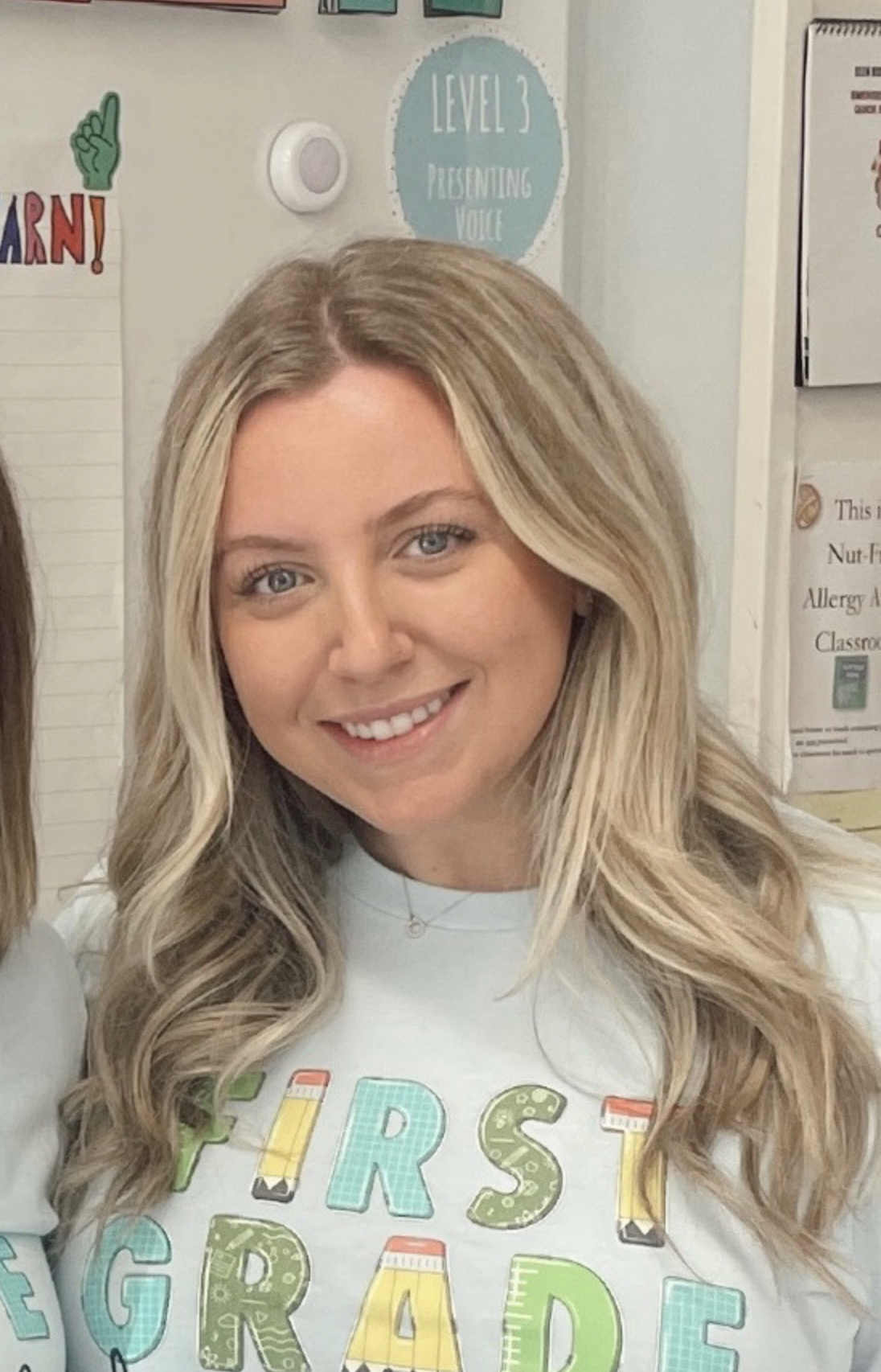 A smiling woman with long, wavy blonde hair and blue eyes wearing a white shirt with colorful block letters and school-themed decorations, indoors in a classroom setting with educational posters and a sign that reads 'LEVEL 3 PRESENTING VOICE' in the background.
