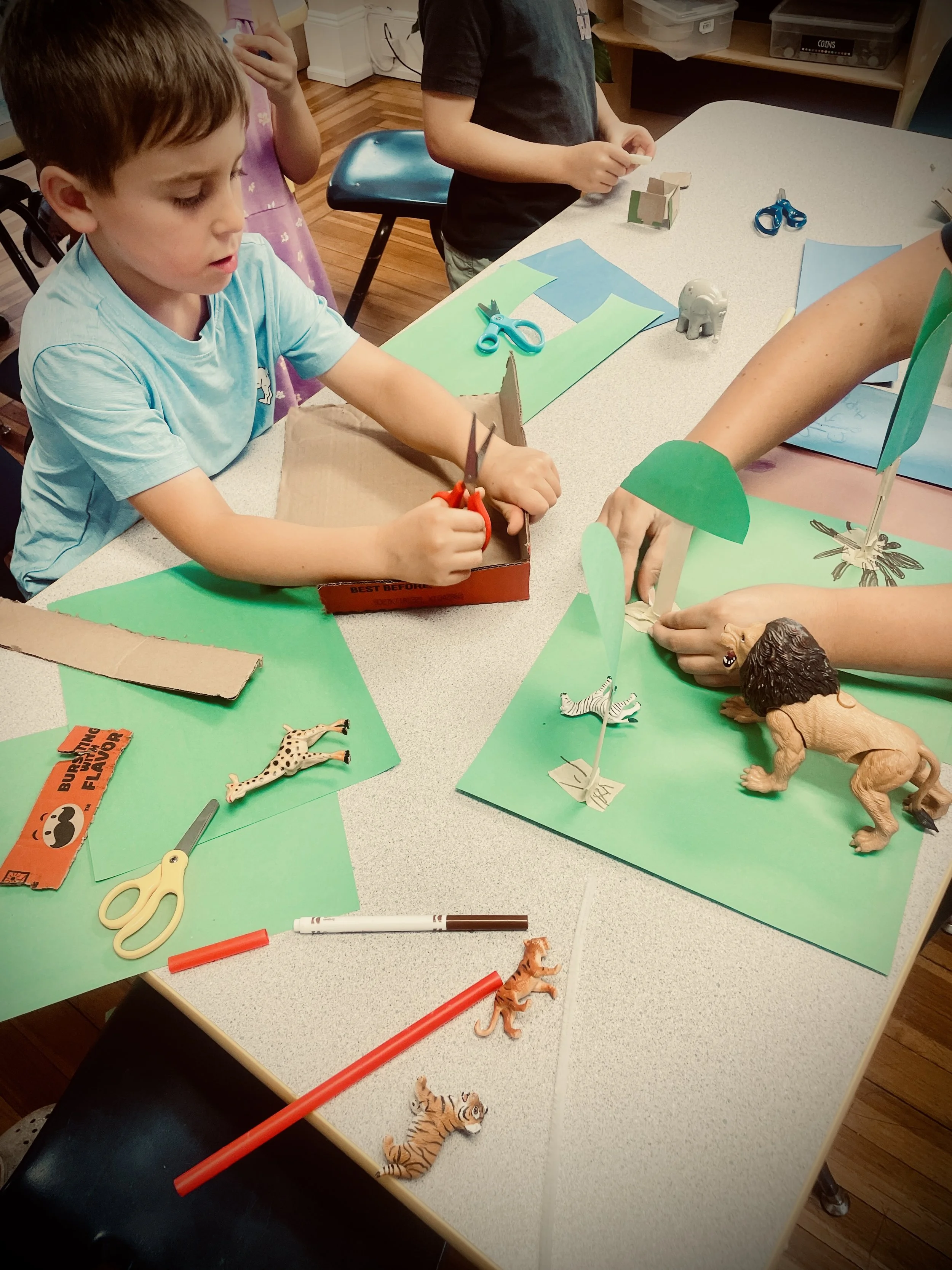 Children engaging in arts and crafts activity with paper, scissors, markers, and toy animals like lions, tigers, elephants, and zebras on a table with colored paper and craft supplies.