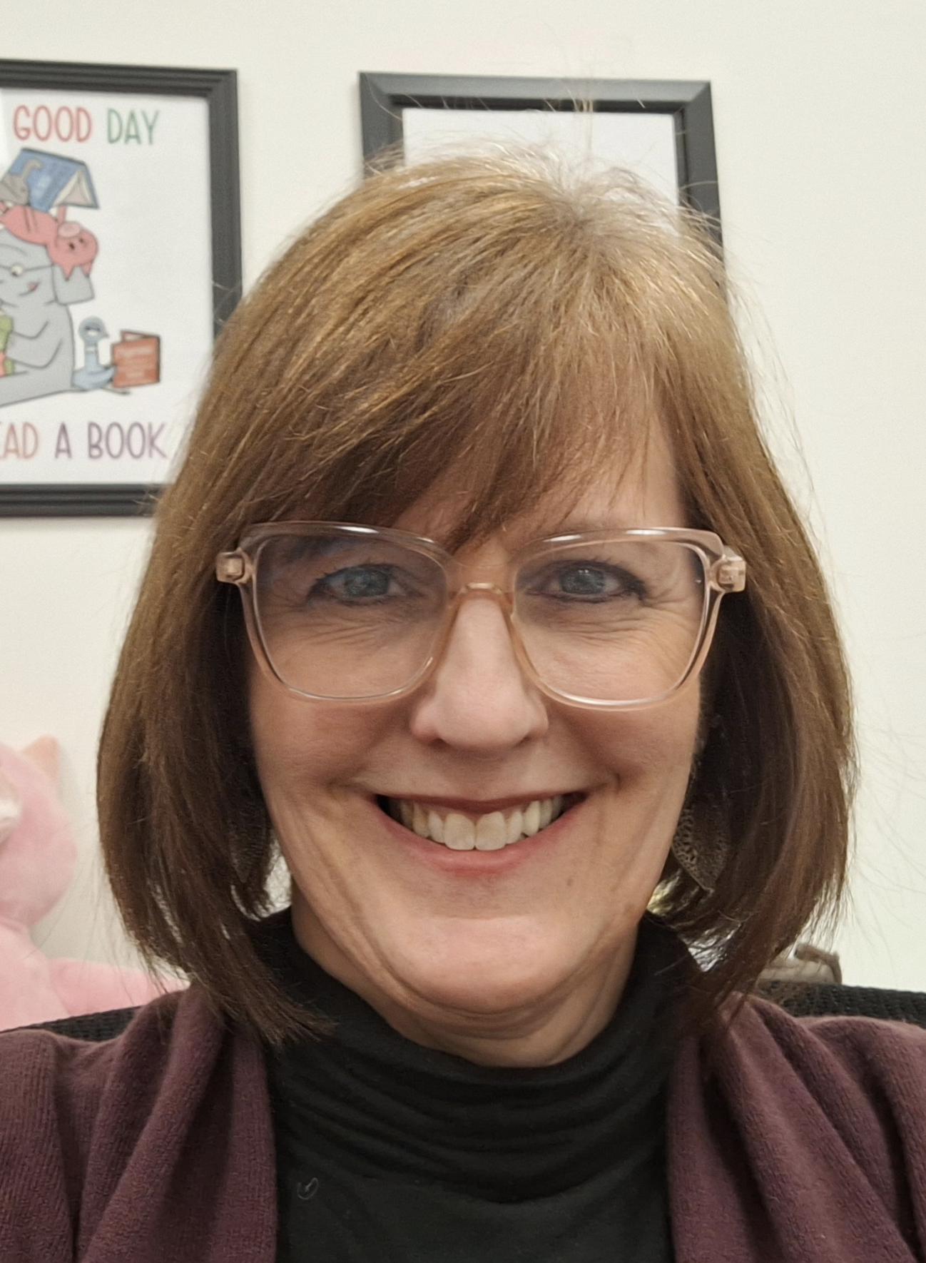 A woman with shoulder-length reddish-brown hair and glasses smiling in an indoor setting, with framed pictures on the wall behind her, including one that says 'Good Day' and shows a cartoon pig reading a book.